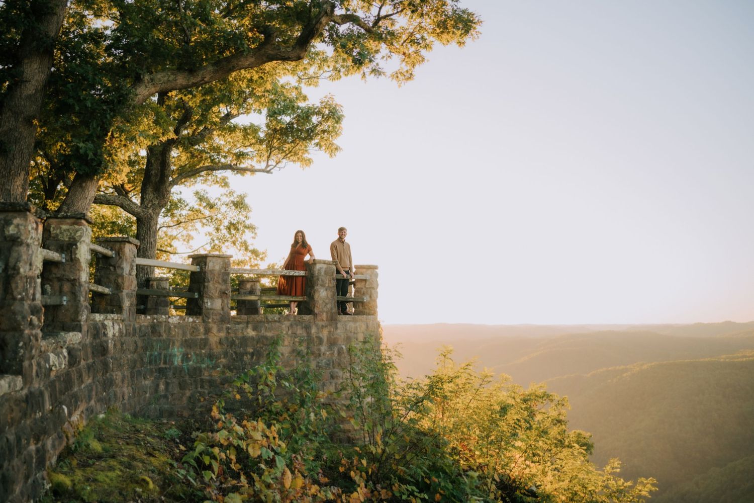 Couple standing on a scenic stone overlook surrounded by lush trees with a sweeping valley view at golden hour.