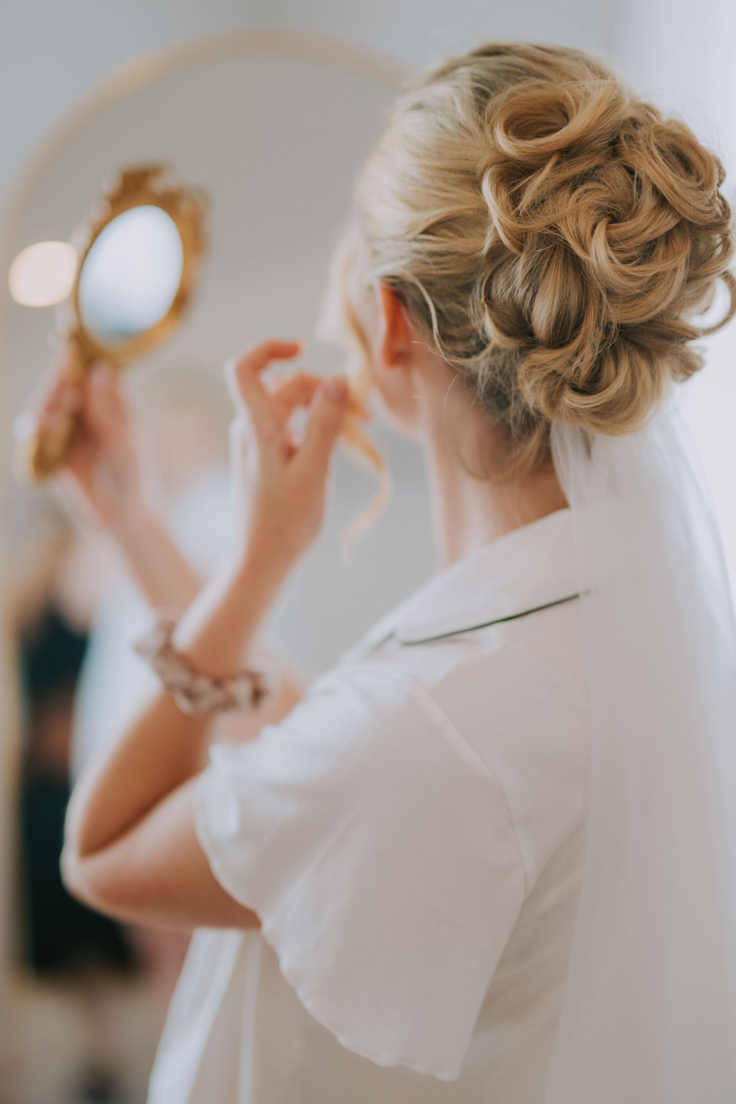 Bride with elegant braided updo and veil holds gold mirror, wearing white robe, preparing for wedding day.