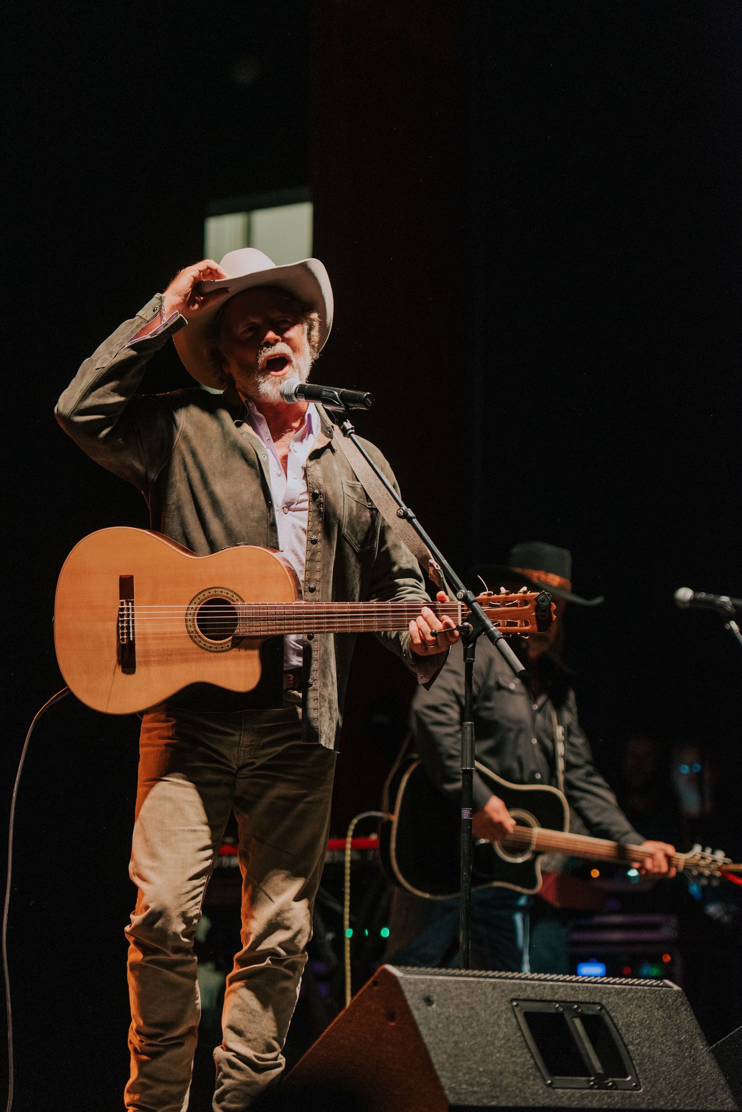 A musician performs on stage with an acoustic guitar while wearing a brown jacket and cowboy hat under stage lighting.