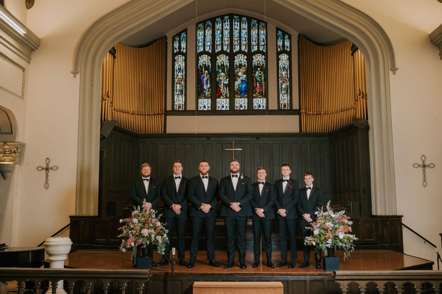 Groomsmen in black tuxedos stand together on stage in front of stained glass windows inside a church.