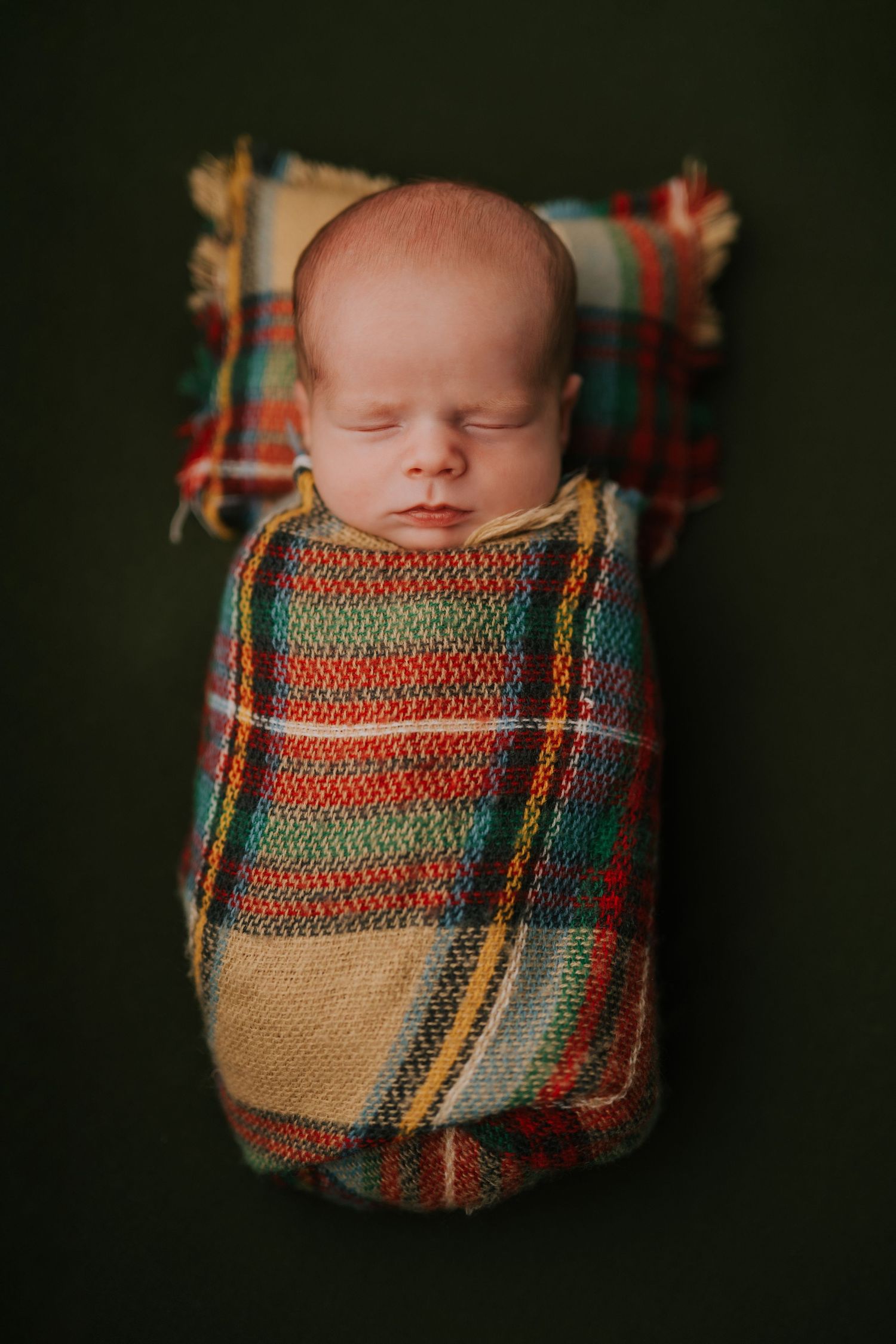 Sleeping newborn wrapped in a colorful plaid blanket against dark background.