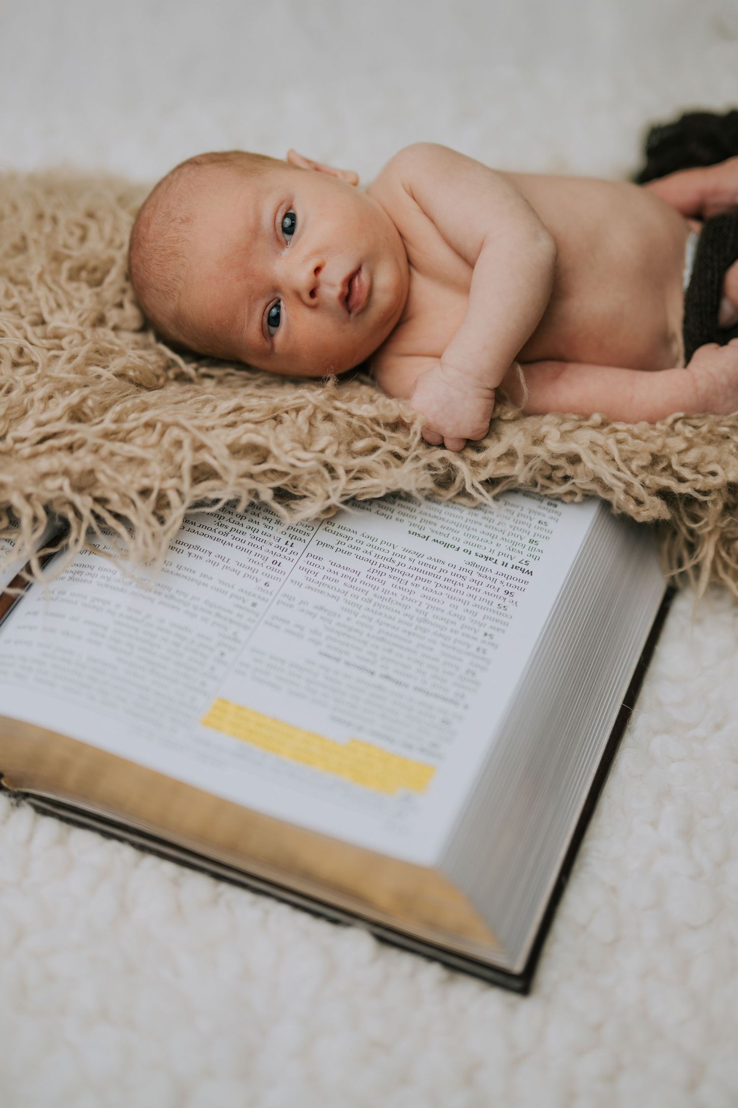 A baby lying on fuzzy carpet over an open book looks directly at camera.