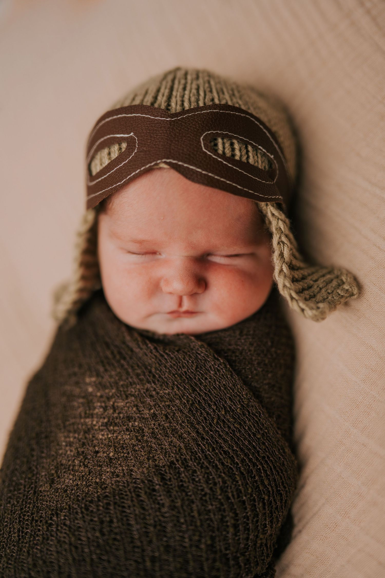 Newborn baby sleeping while wearing knit aviator cap on textured backdrop.