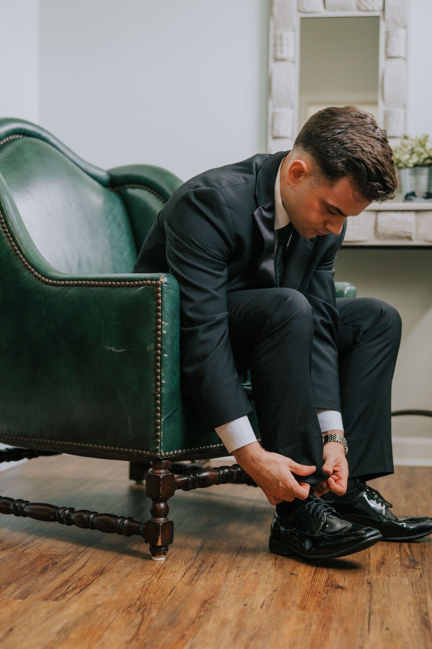 Person in black suit putting on dress shoes while sitting on green leather chair.