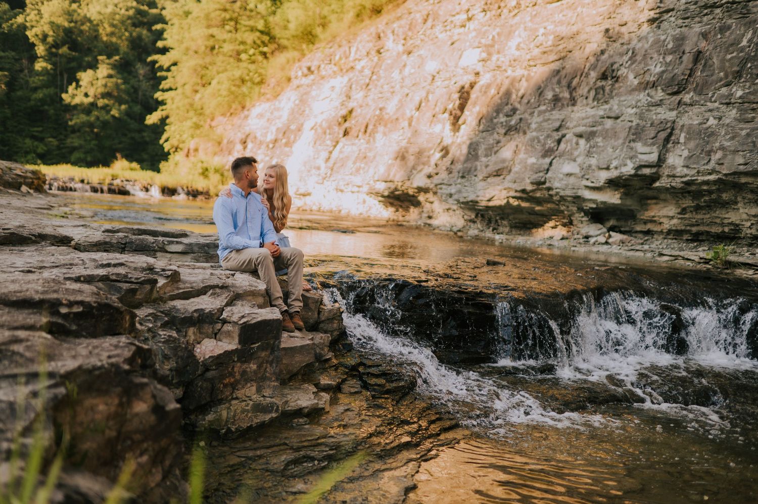 A couple sits together on rocky terrain near a small waterfall during golden hour.