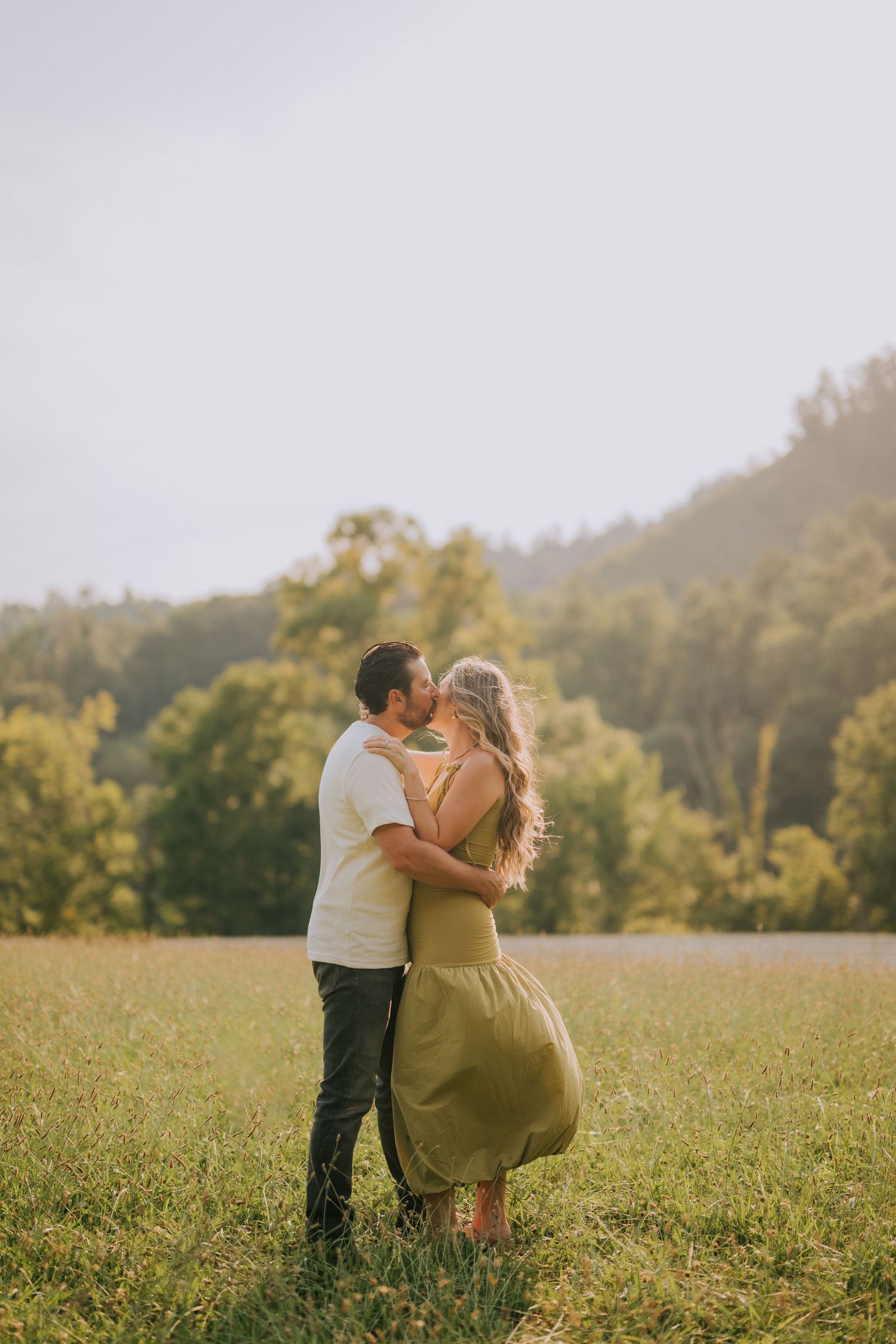 Romantic moment captured in a sunlit field with mountains and trees in the distance.