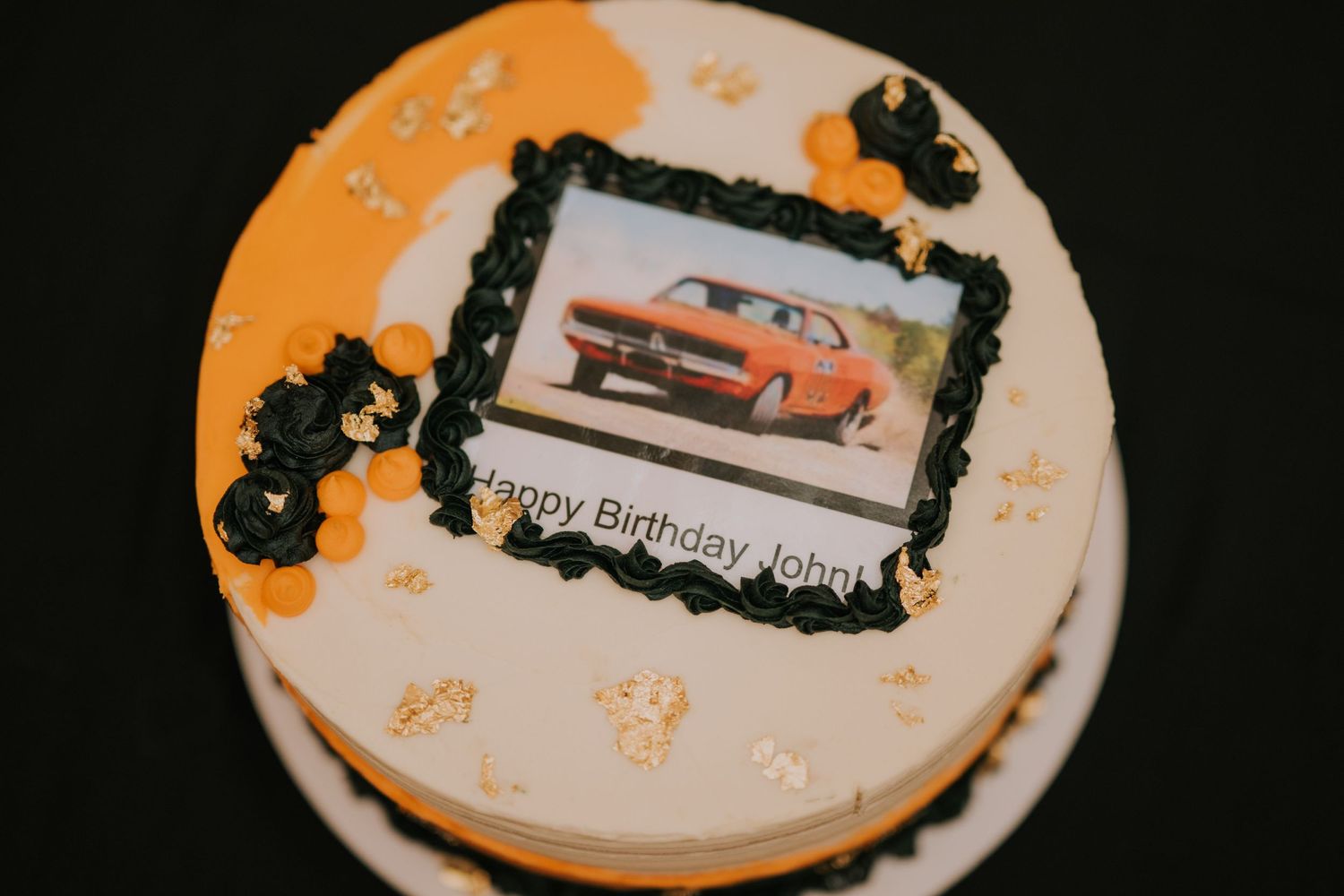 A round white and orange birthday cake decorated with a vintage orange car photo and black floral accents.