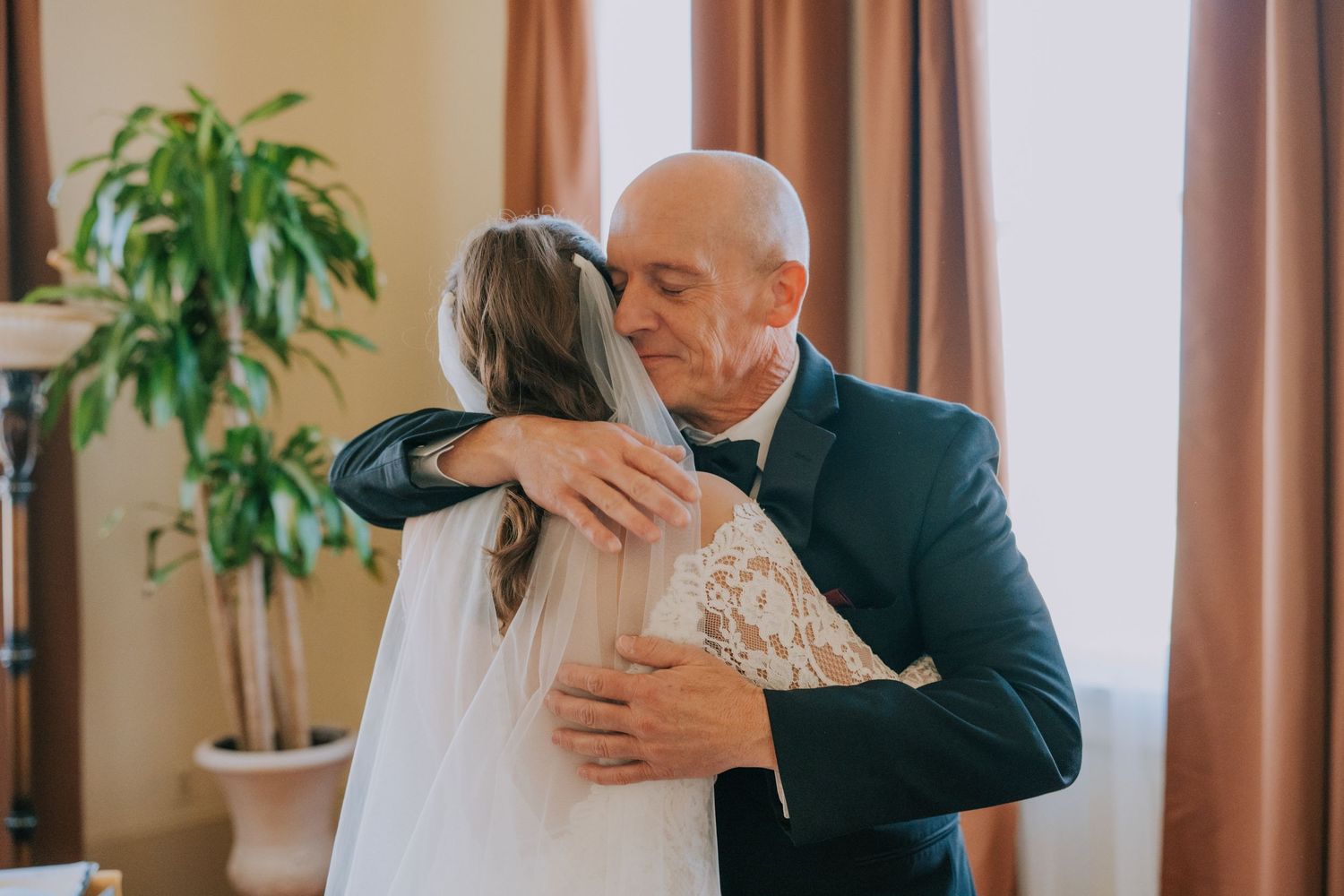 An emotional moment as bride and man embrace during a wedding day near brown curtains and houseplant.