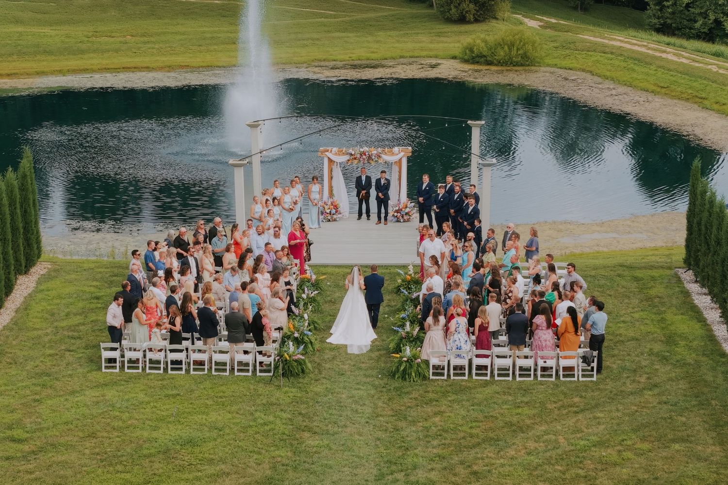Outdoor wedding ceremony by a pond with fountain, guests seated in white chairs on green lawn under sunny skies.