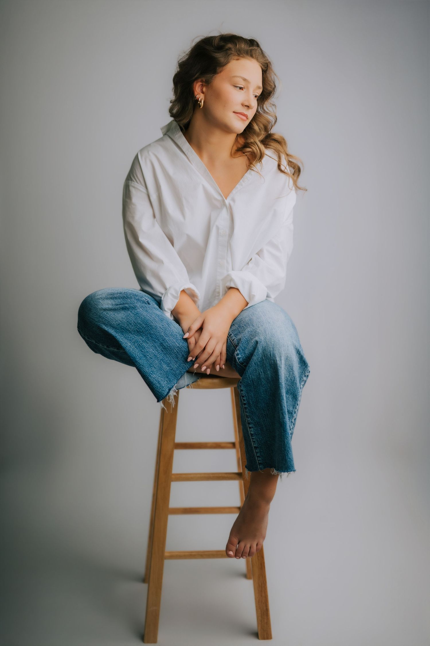 Young woman in white shirt and blue jeans sitting barefoot on wooden stool against gray background.