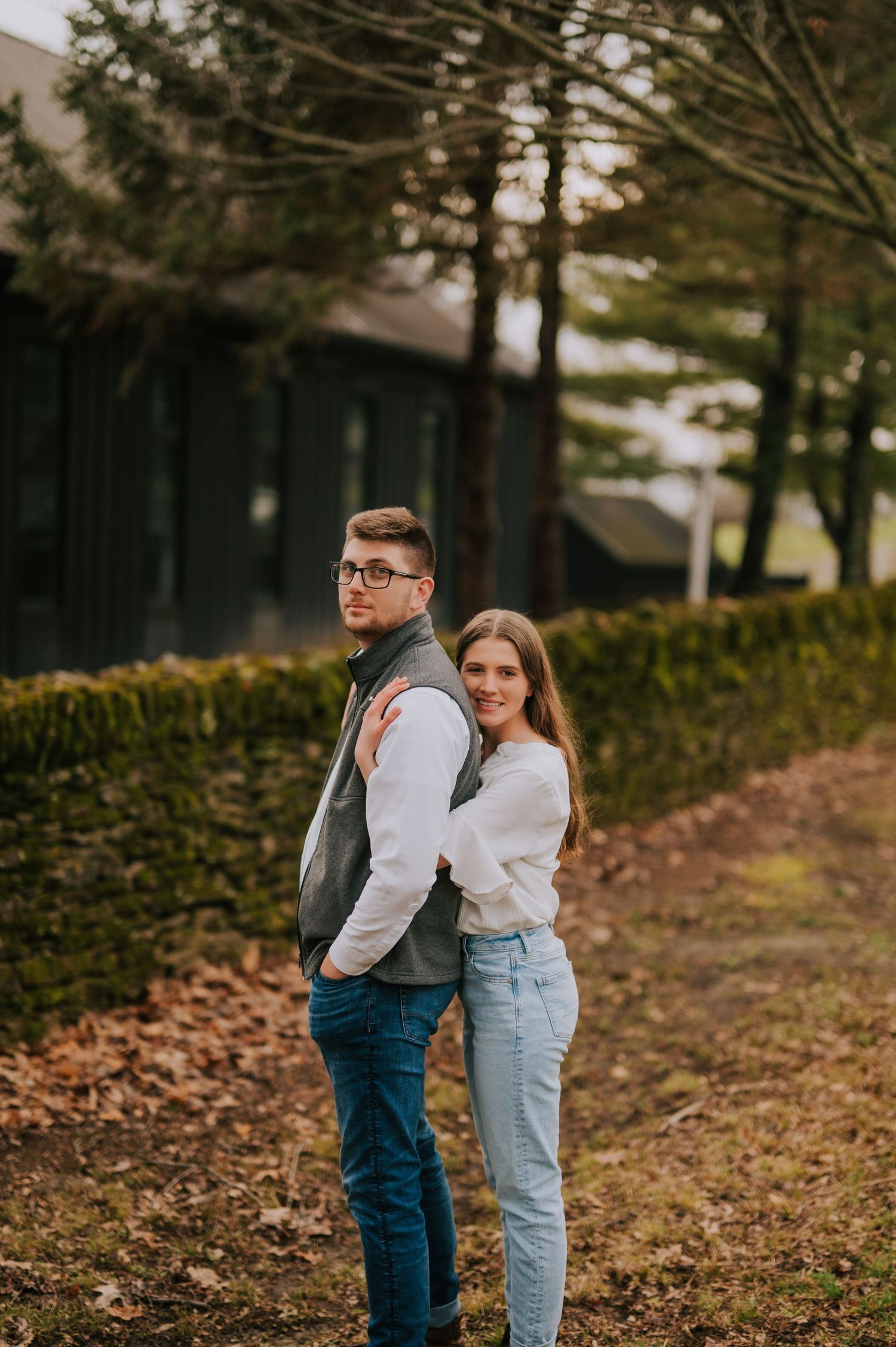 A couple poses together among fallen leaves and hedges during an autumn photoshoot.