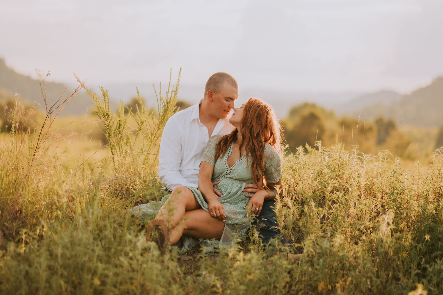 A couple embraces in a sunlit field with mountains in the background.