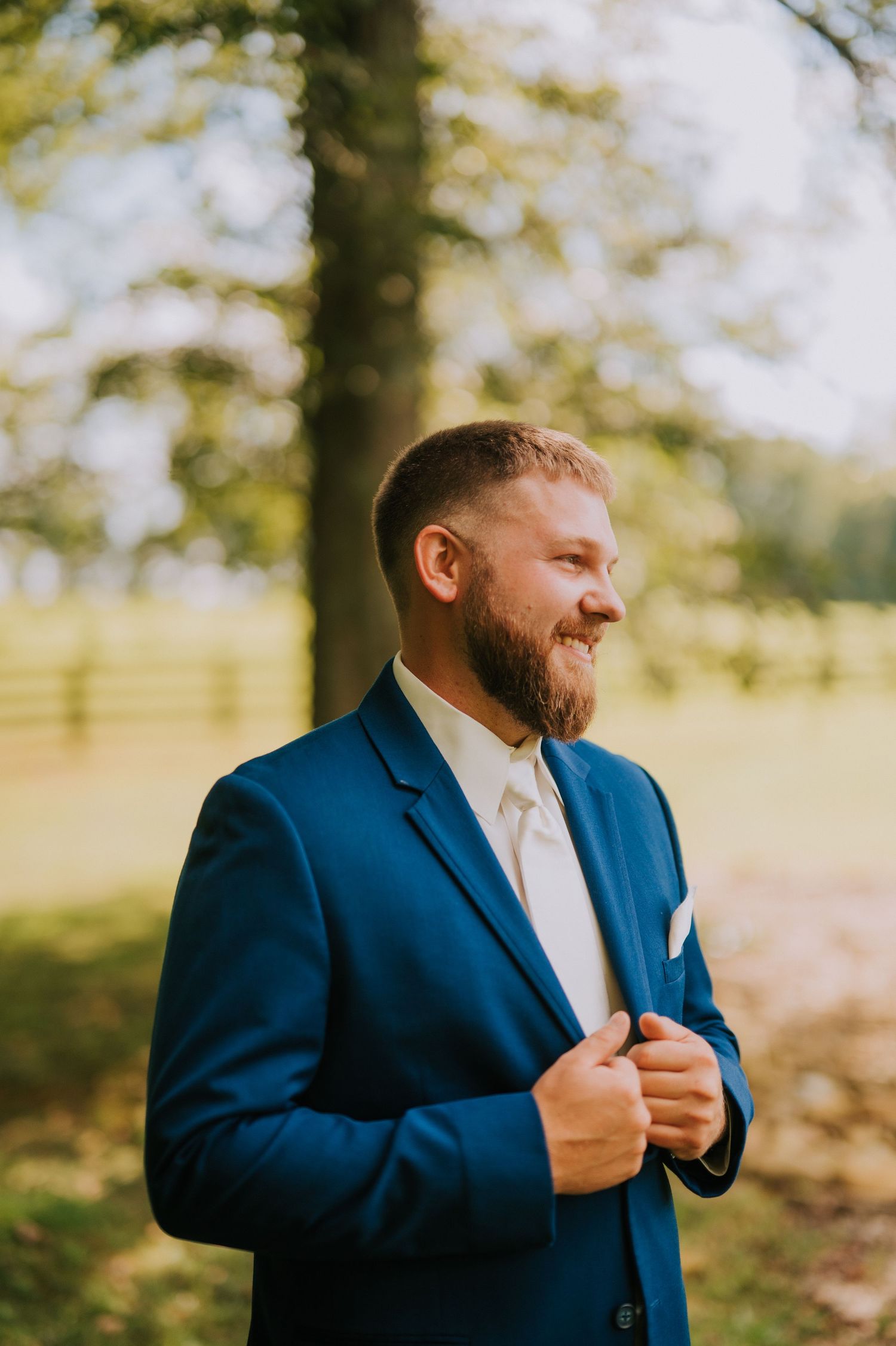 A groom in a navy blue suit and white tie adjusts his jacket outdoors on his wedding day.