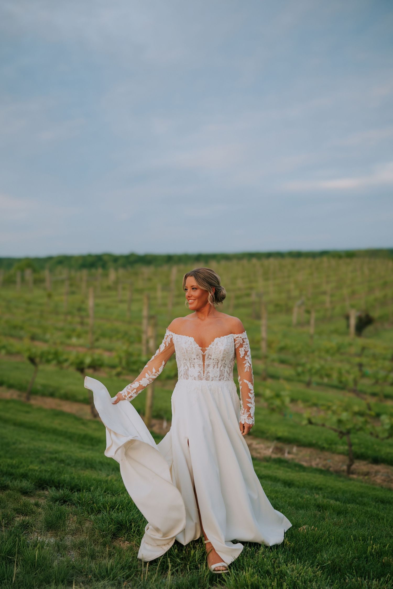 Bride in flowing lace gown twirling in vineyard setting during sunset.