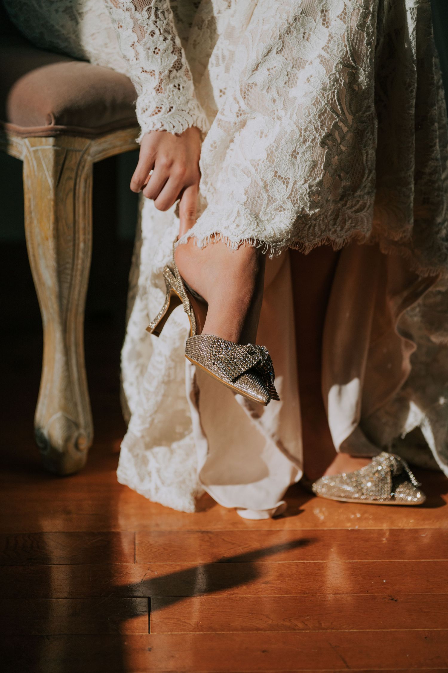 Close-up of a bride's leg showing a sparkly wedding shoe and white lace dress hem against a wooden floor.