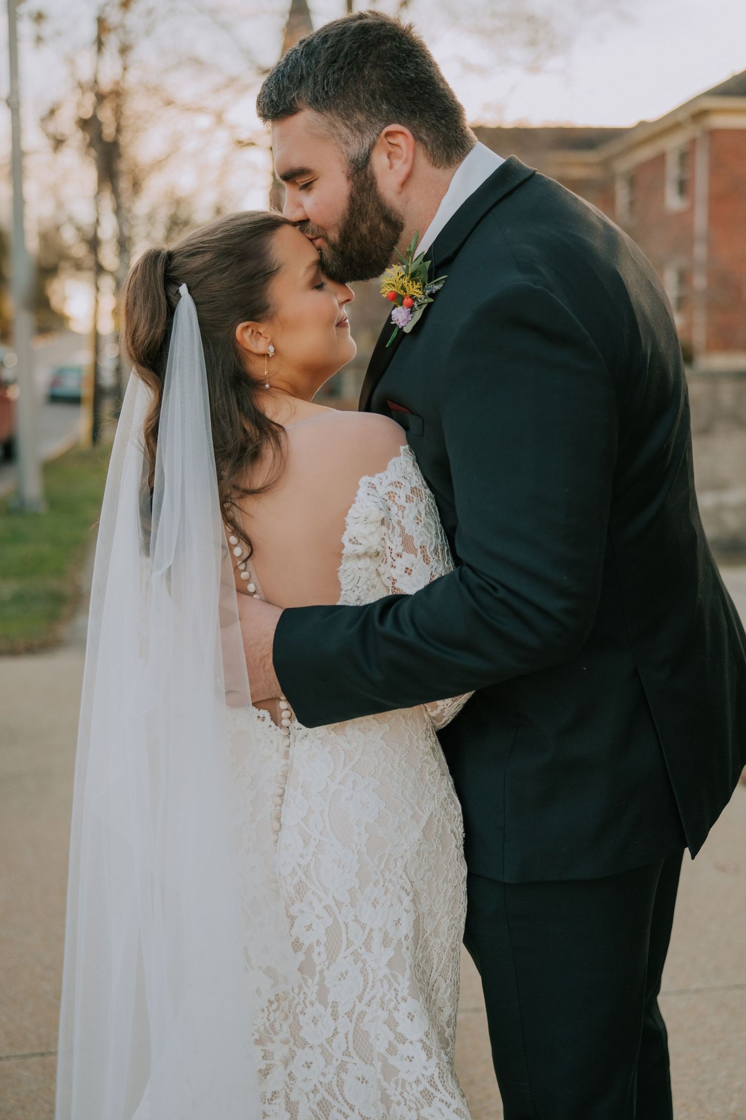A tender moment between newlyweds sharing an intimate kiss outdoors, with the bride in lace and groom in classic suit.