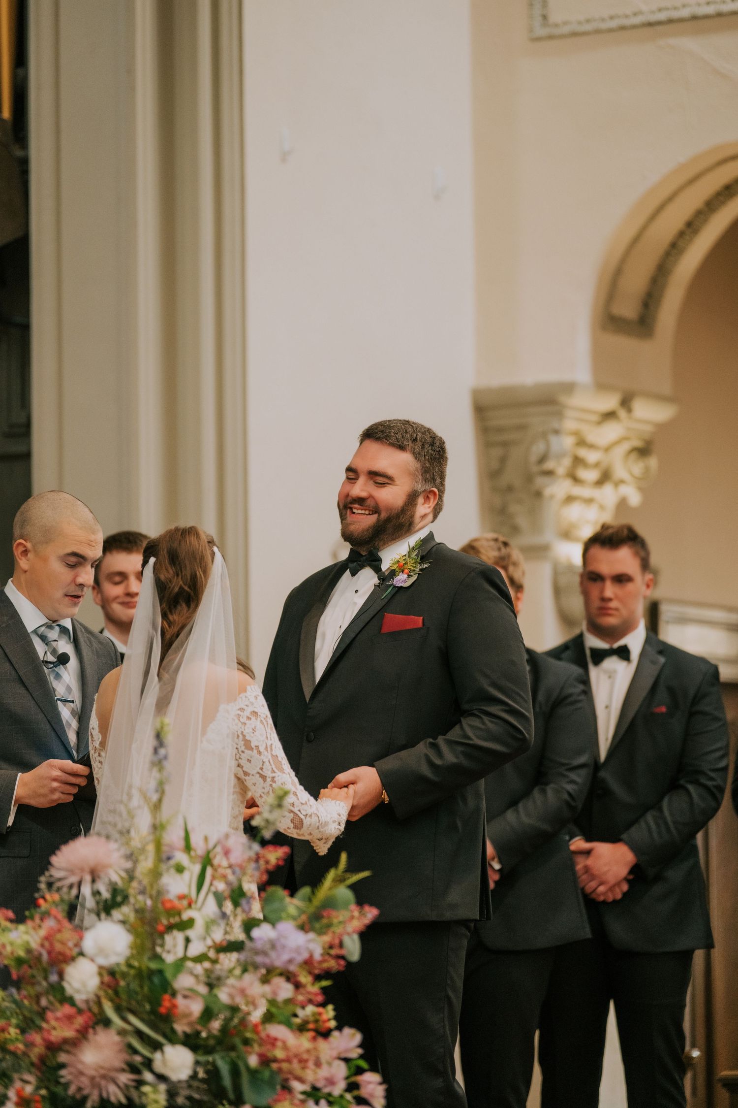 Wedding ceremony in ornate church interior with floral arrangements and attendants at the altar.