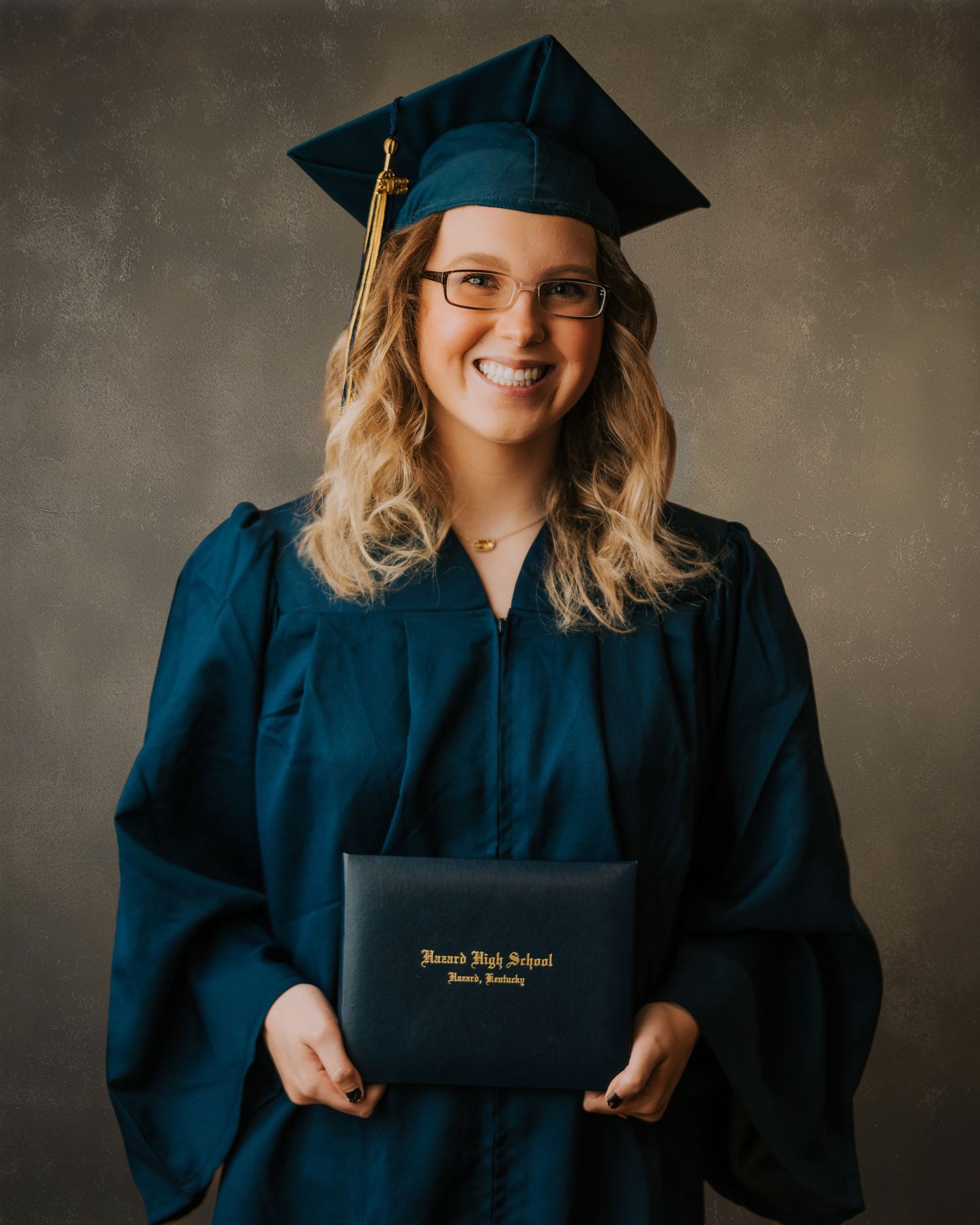 Smiling graduate in teal cap and gown holding Howard High School diploma during professional portrait session.