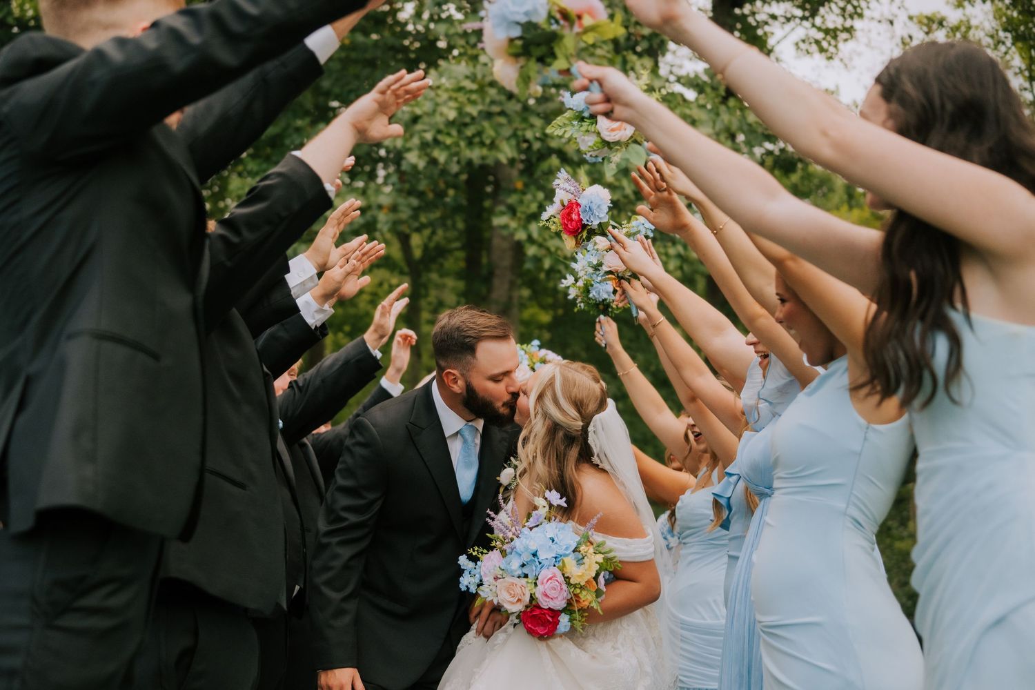 Bride and groom kissing under raised hands arch formed by wedding party outdoors, surrounded by colorful bouquets.