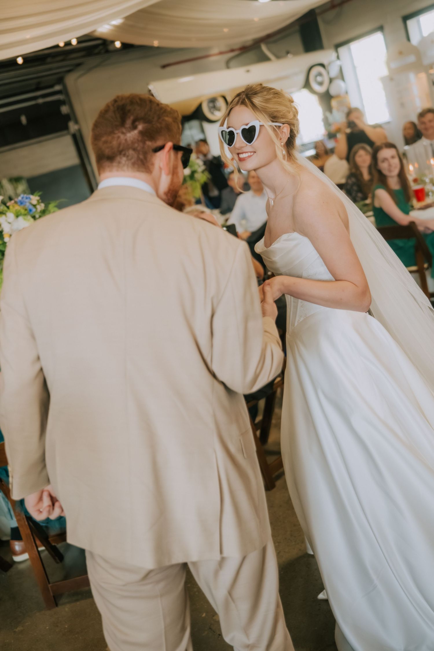 Bride wearing heart-shaped sunglasses holds hands with groom in tan suit during indoor wedding ceremony.