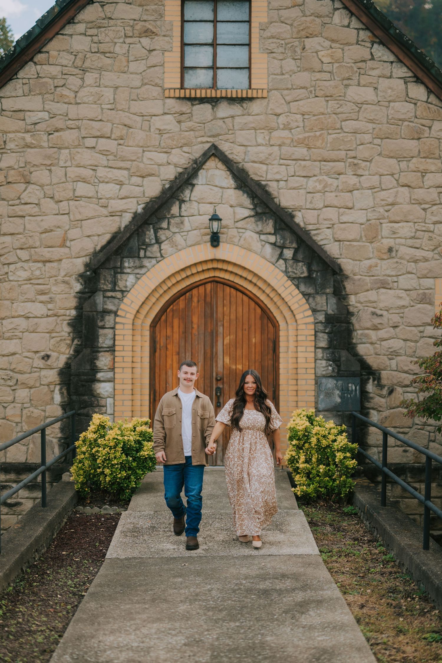 A couple stands in front of a beautiful stone church with arched wooden doors.