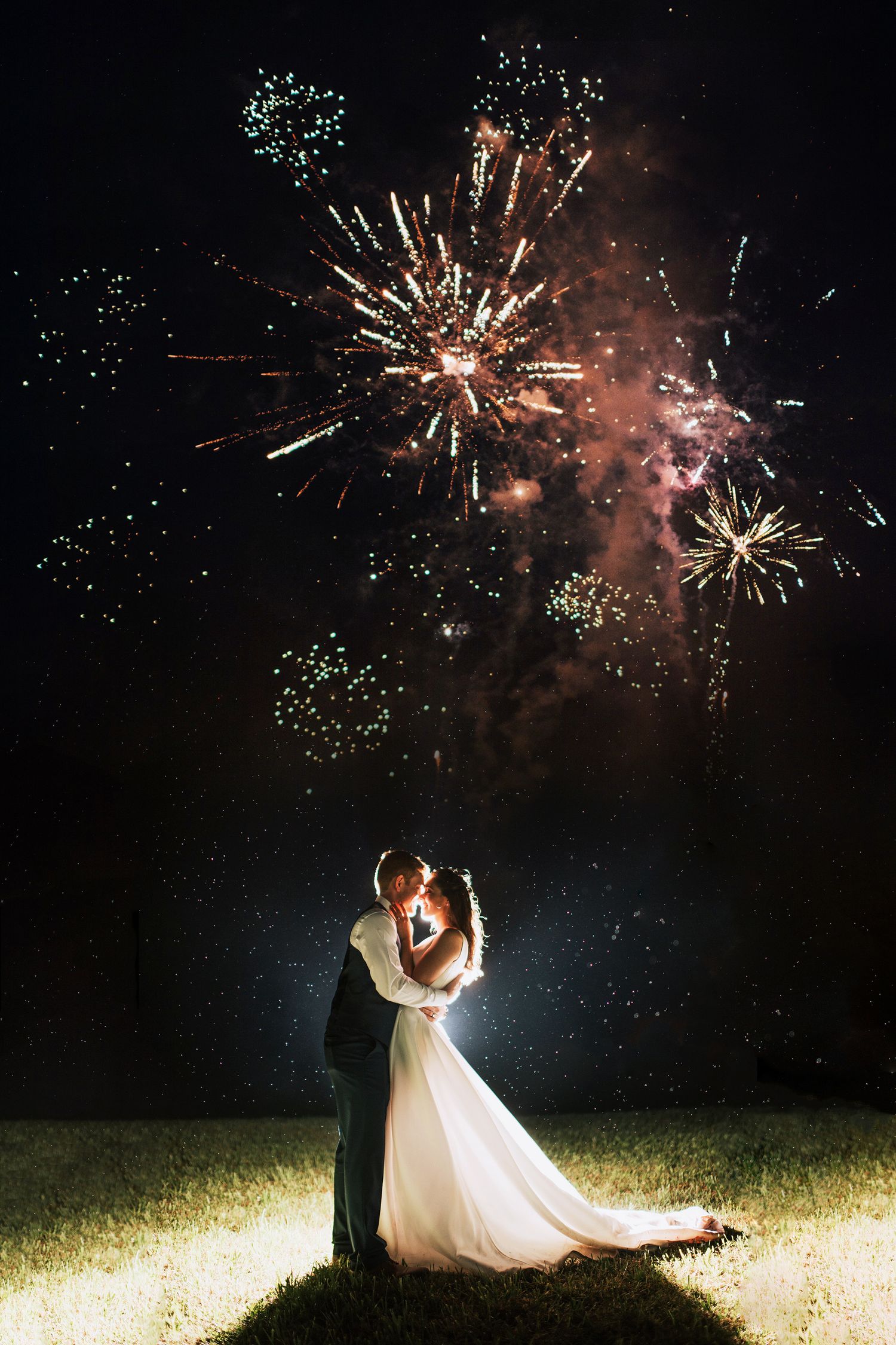 A couple shares a romantic moment under bursting fireworks in the night sky during their wedding celebration.