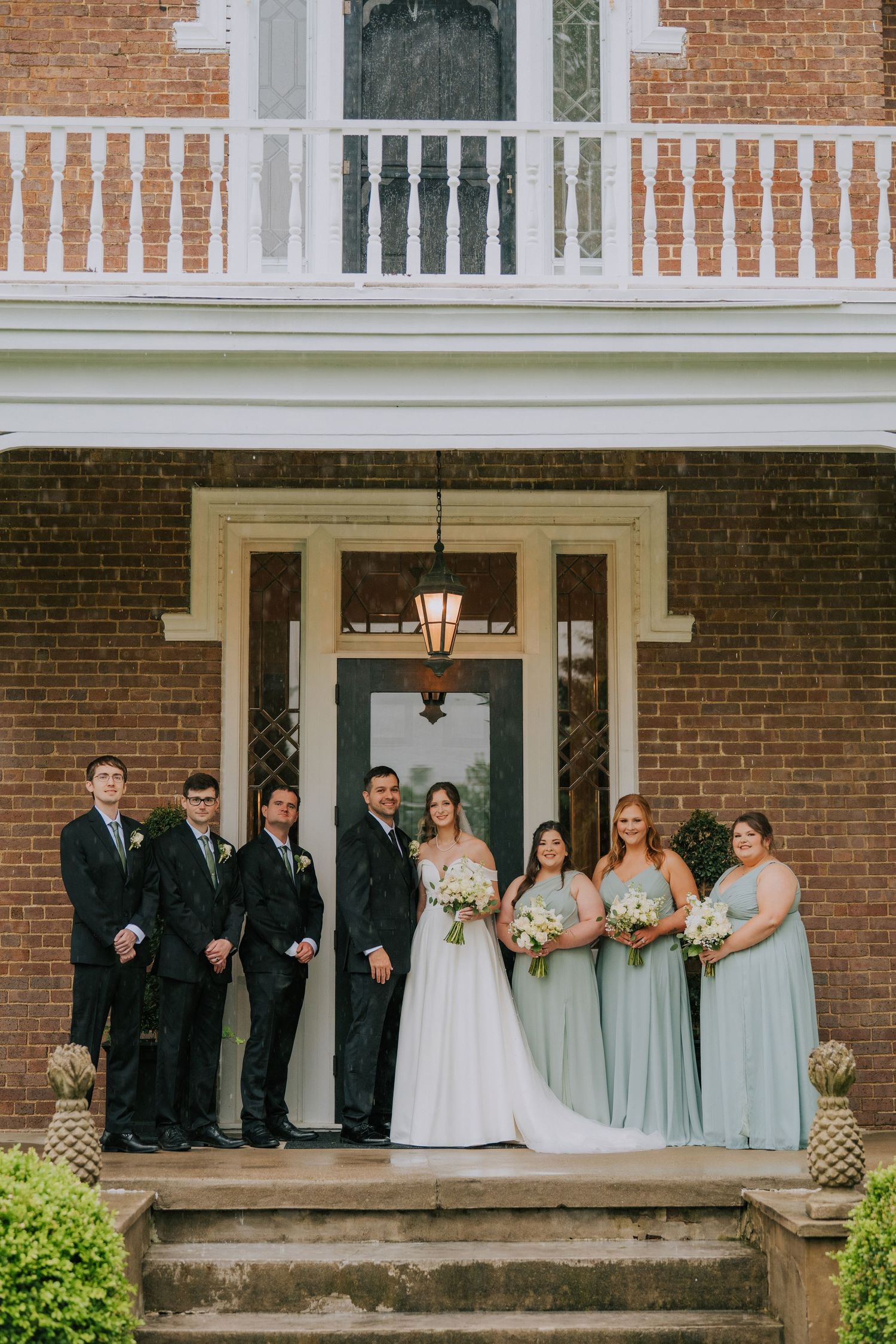 Wedding party poses on a classic brick mansion porch in formal attire.
