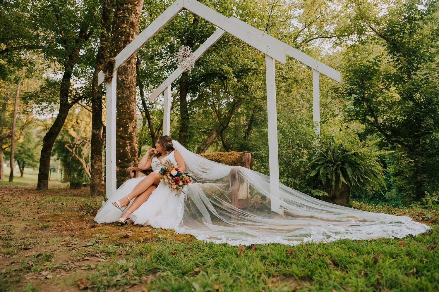 Bride lying gracefully under white wedding arch outdoors with flowing veil and train.