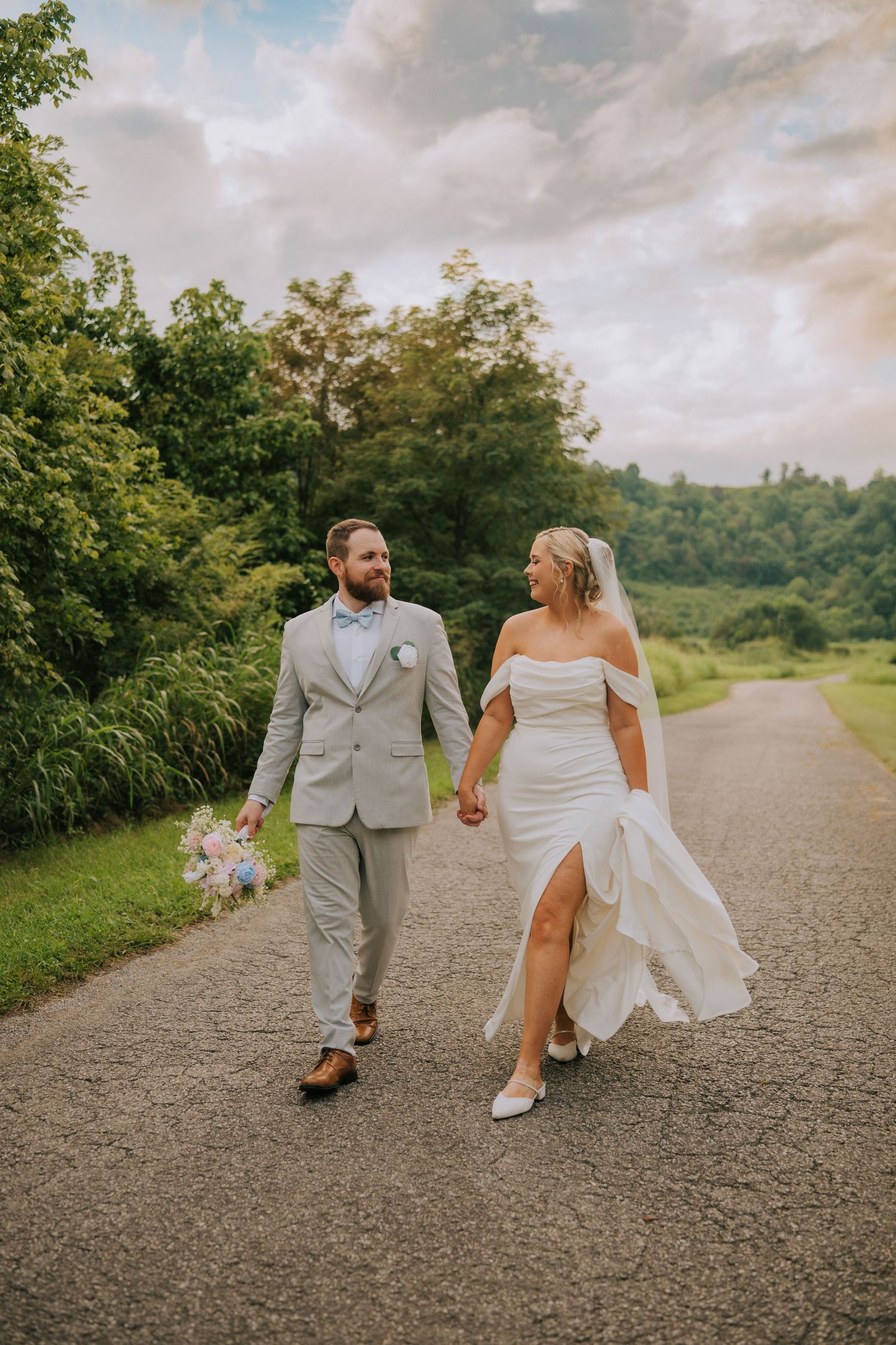 A bride and groom walk hand in hand down a gravel path surrounded by lush greenery.