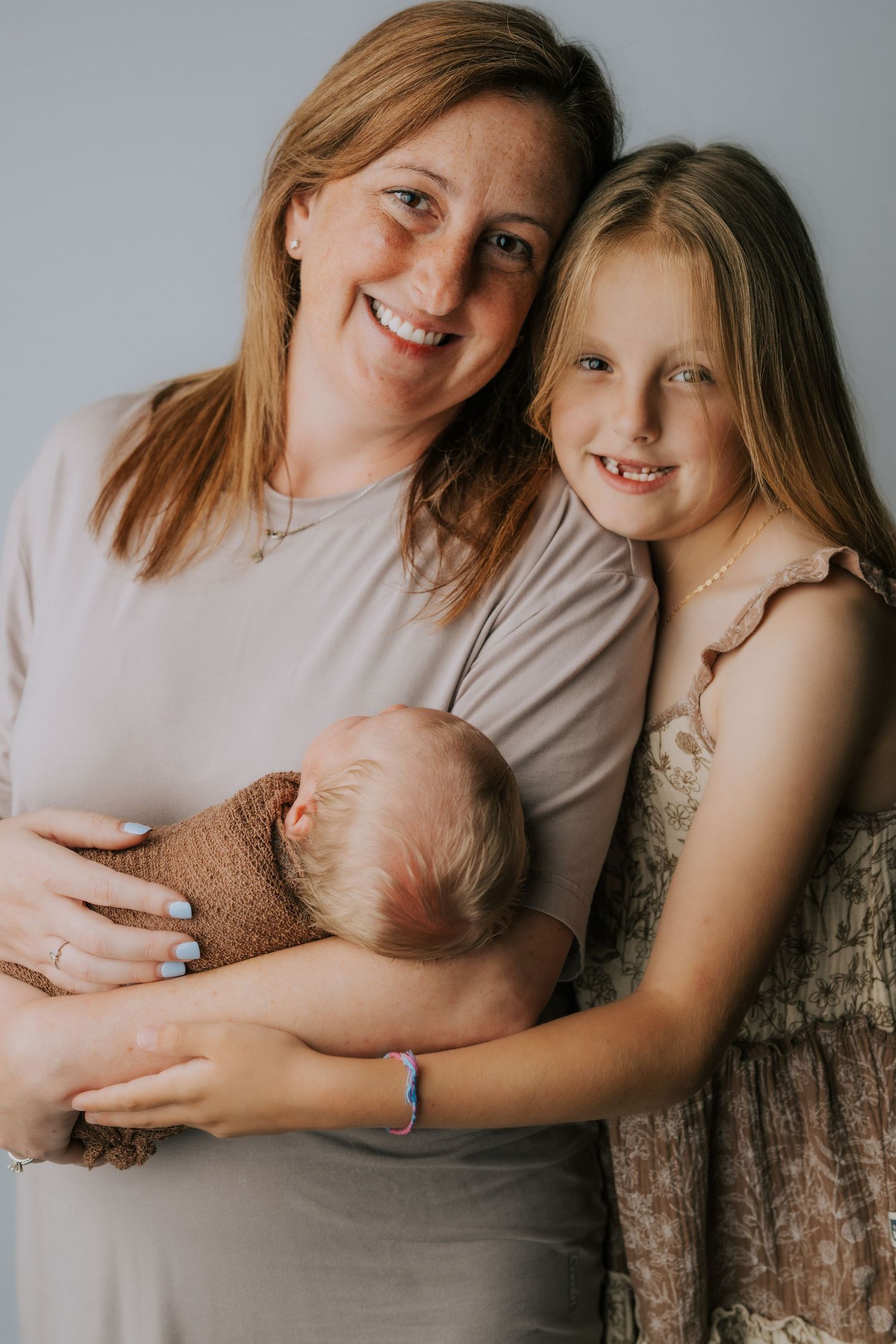 A smiling mother holds a newborn baby wrapped in brown while an older blonde girl hugs them both.