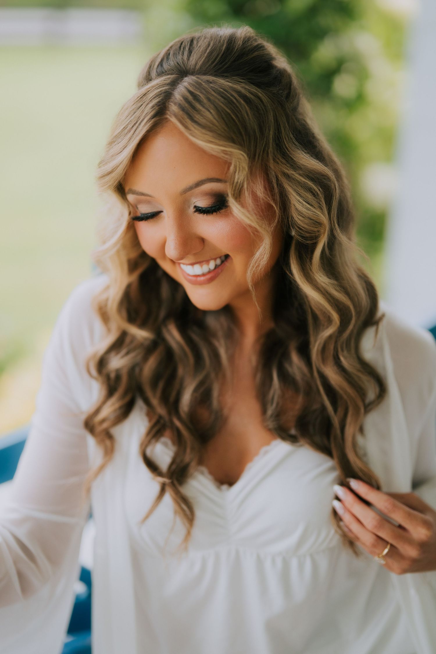 Smiling bride with long wavy hair in half-up style wearing white dress, looking down elegantly before wedding ceremony.