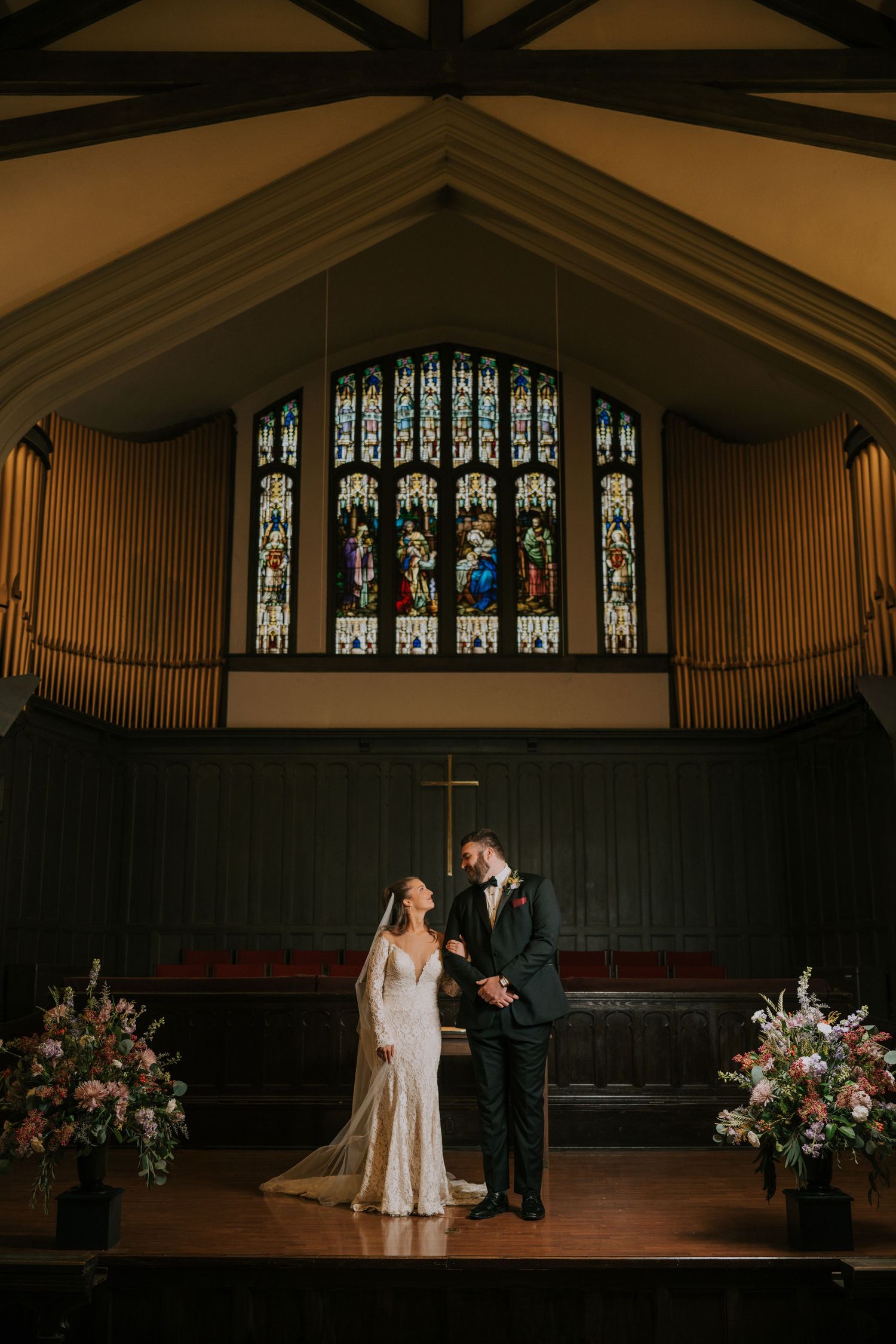 Elegant church wedding ceremony with stained glass windows, organ pipes, and floral arrangements in warm ambient lighting.