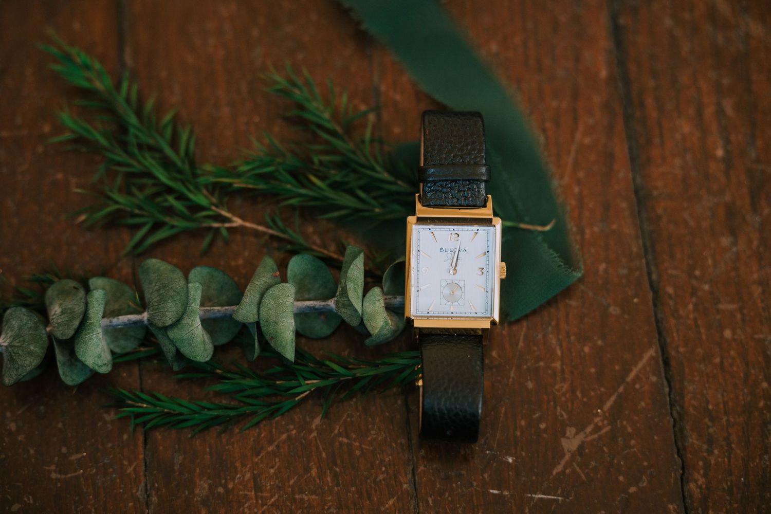 Elegant gold wristwatch with black leather strap displayed on wooden surface with fresh eucalyptus sprigs.