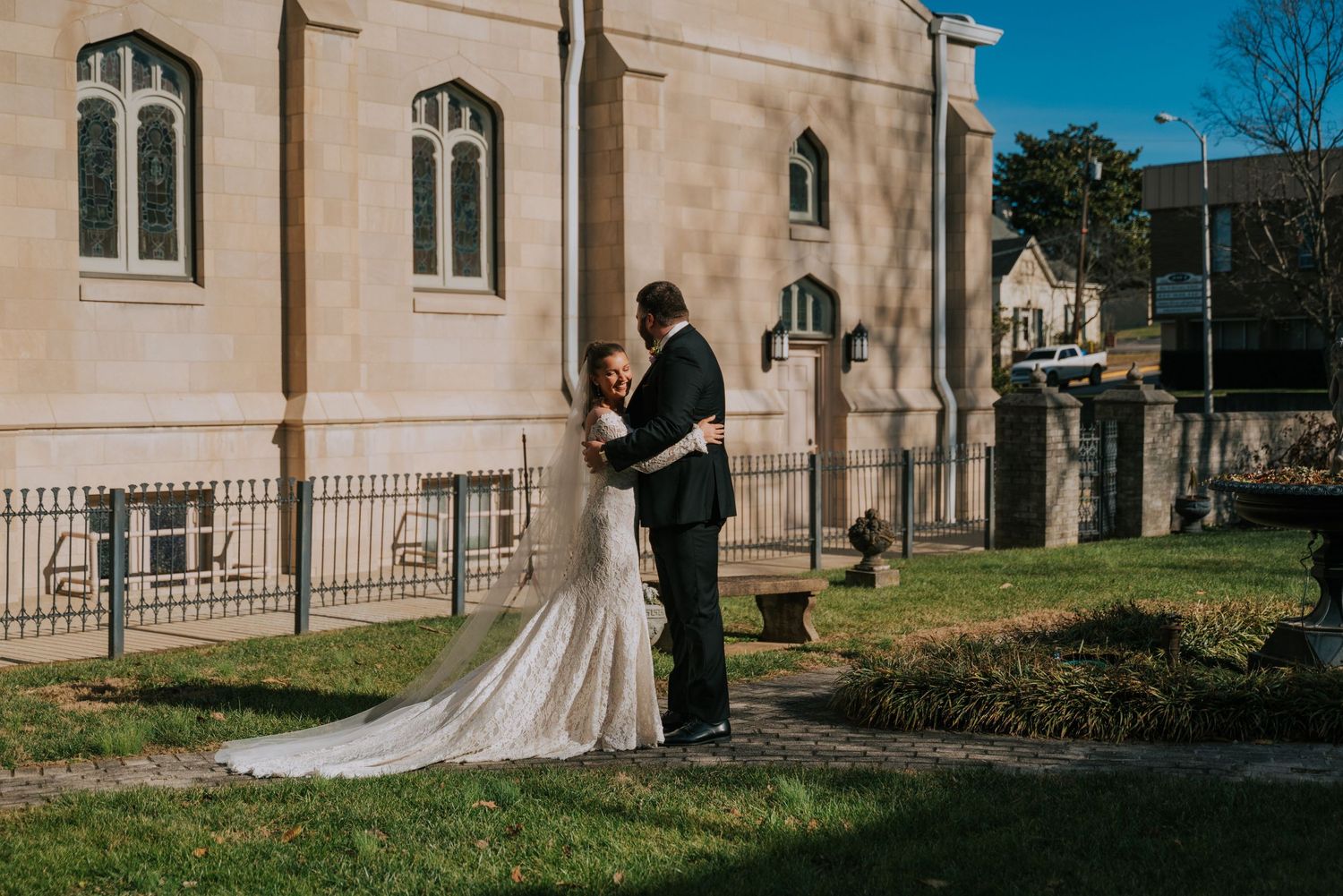 A couple embraces during their wedding photoshoot outside a historic stone church with arched windows on a sunny day.