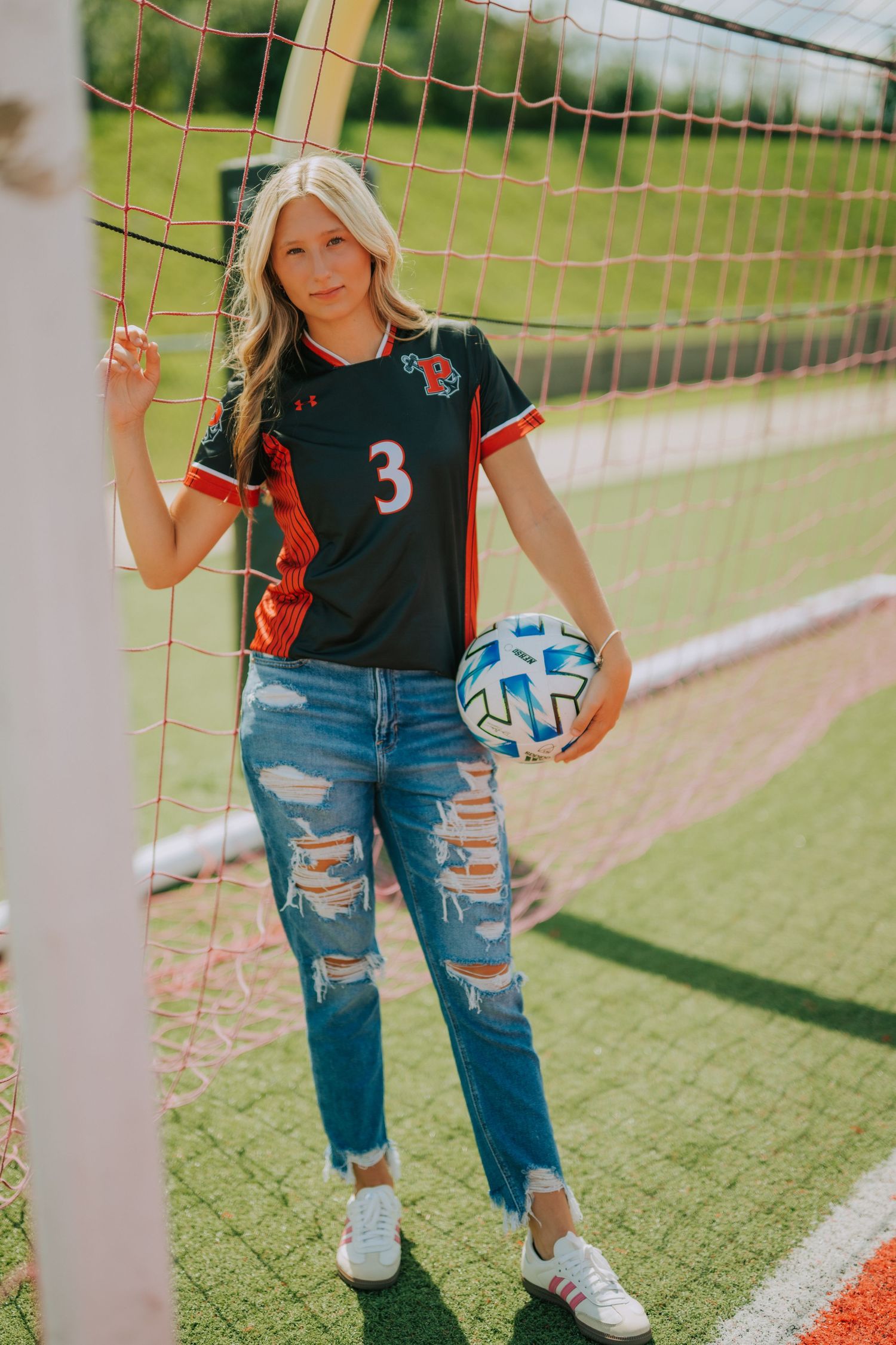 Student athlete poses with volleyball on outdoor court wearing ripped jeans and jersey.