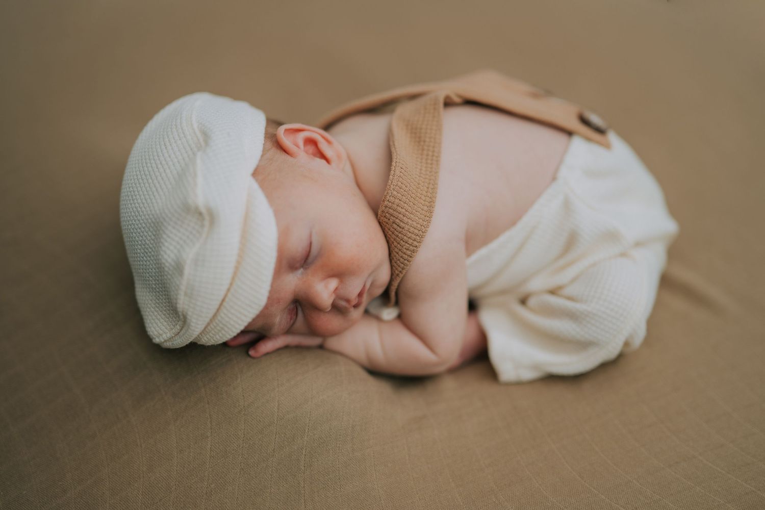 A peaceful sleeping newborn in beige outfit and white cap on brown backdrop.