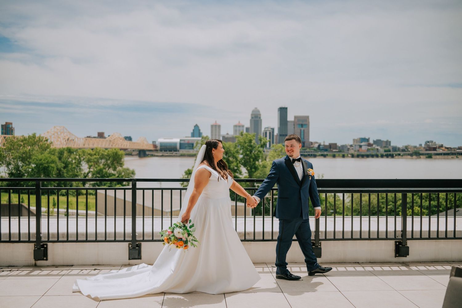 Wedding couple poses along riverside with city skyline in background.