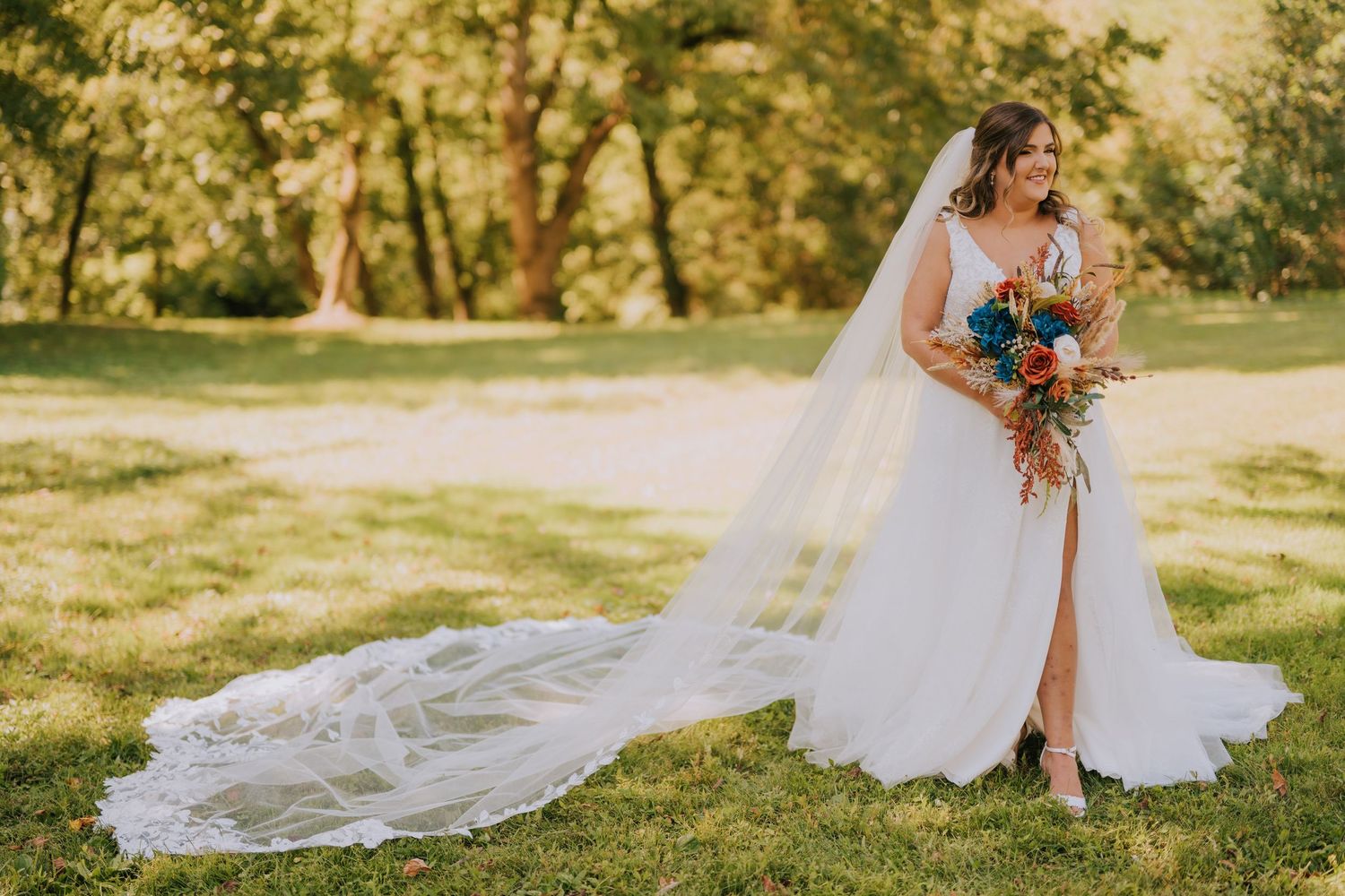 A flowing white wedding veil trails behind on grass in golden sunlight during an outdoor wedding ceremony.