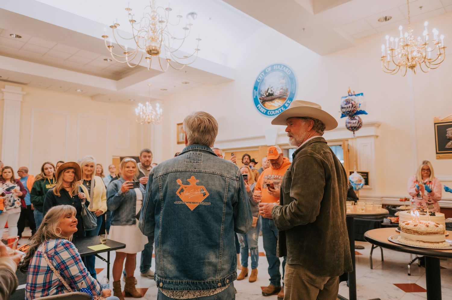 A group of people gather at an indoor event with chandeliers while someone wearing a cowboy hat speaks to the crowd.