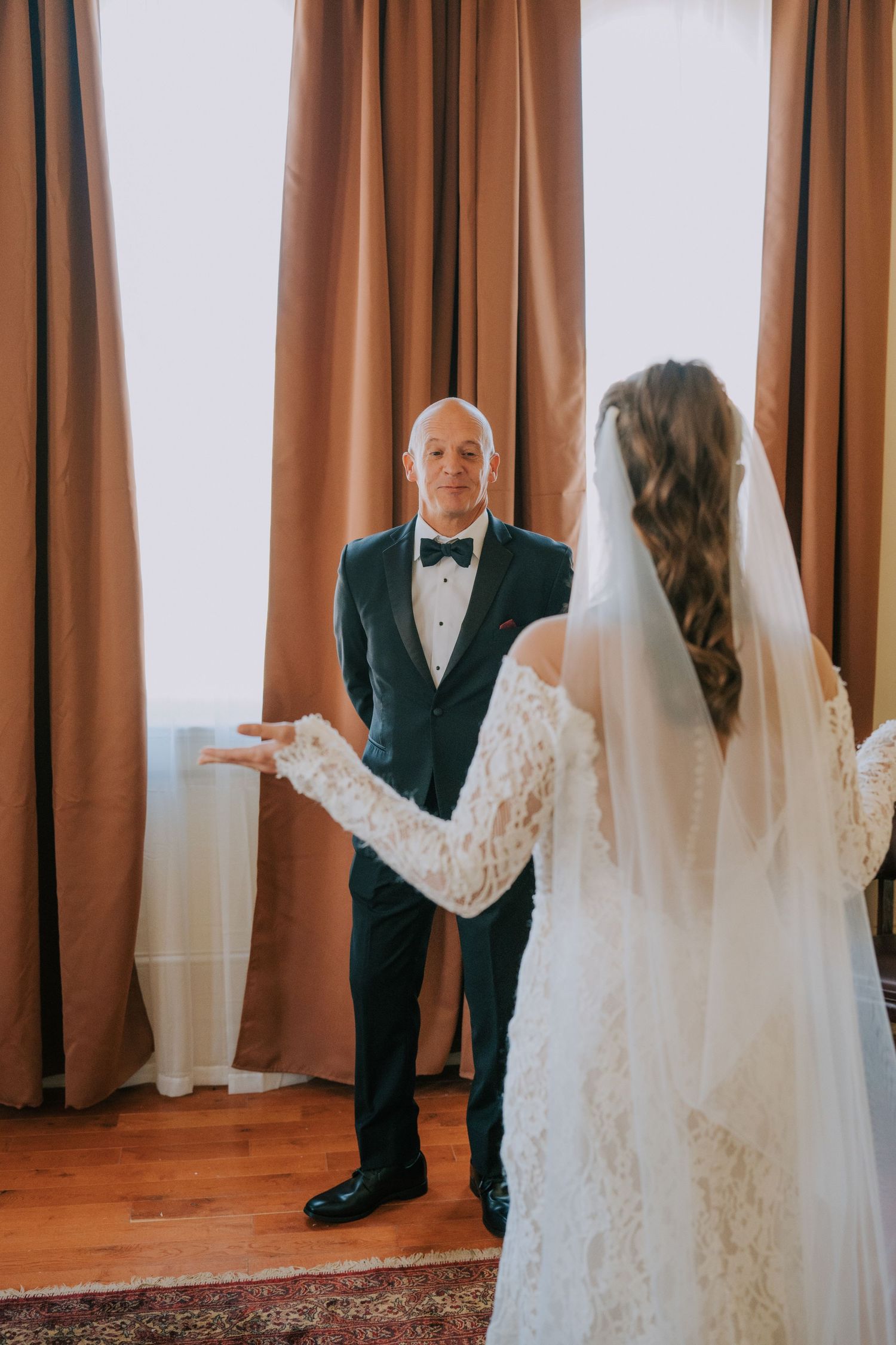 A bride in lace dress and veil shares a moment with a man in tuxedo by brown curtains.