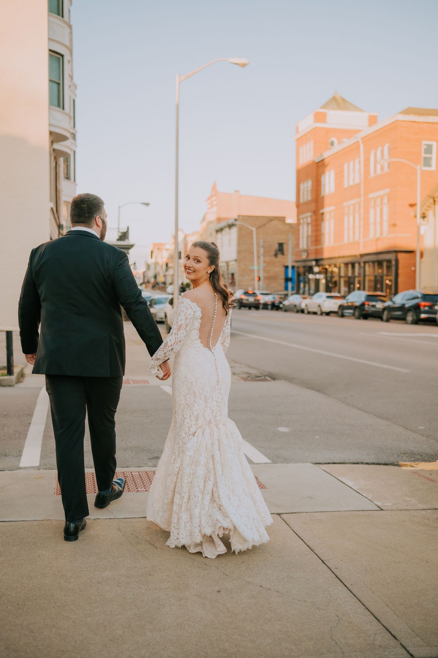 A couple in wedding attire walks down a city sidewalk with vintage brick buildings in the background.