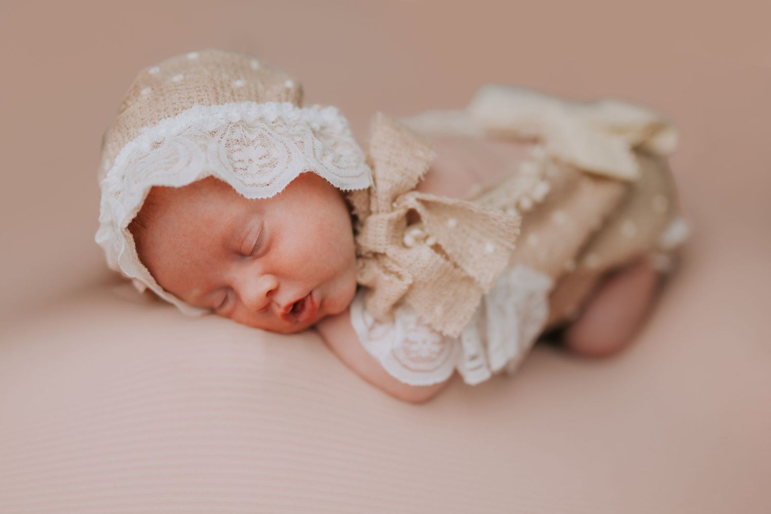 Sleeping newborn baby in lace bonnet and ruffled outfit on beige background.
