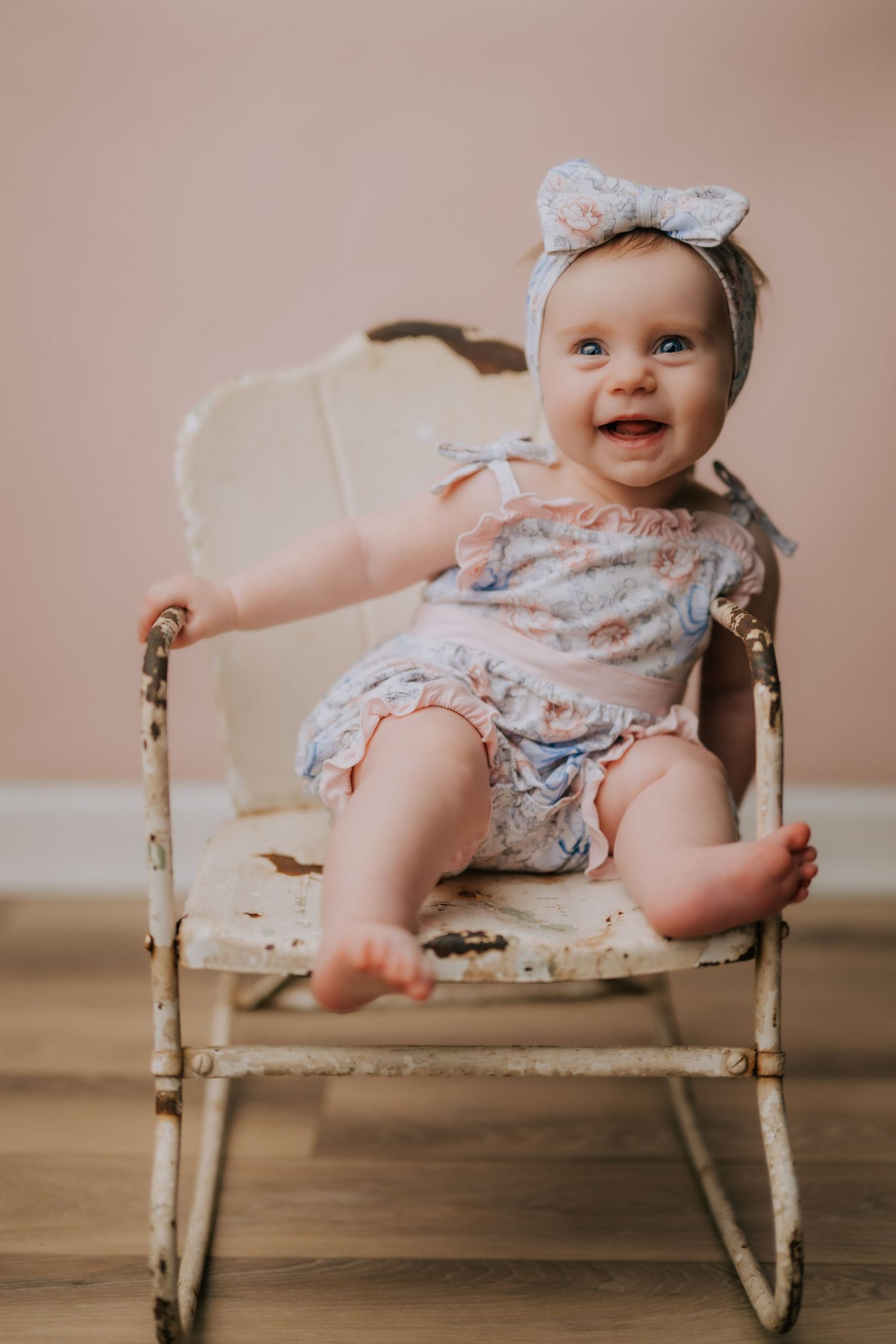 Smiling baby girl in floral romper and bow headband sitting in a vintage white rocking chair against a pink wall.