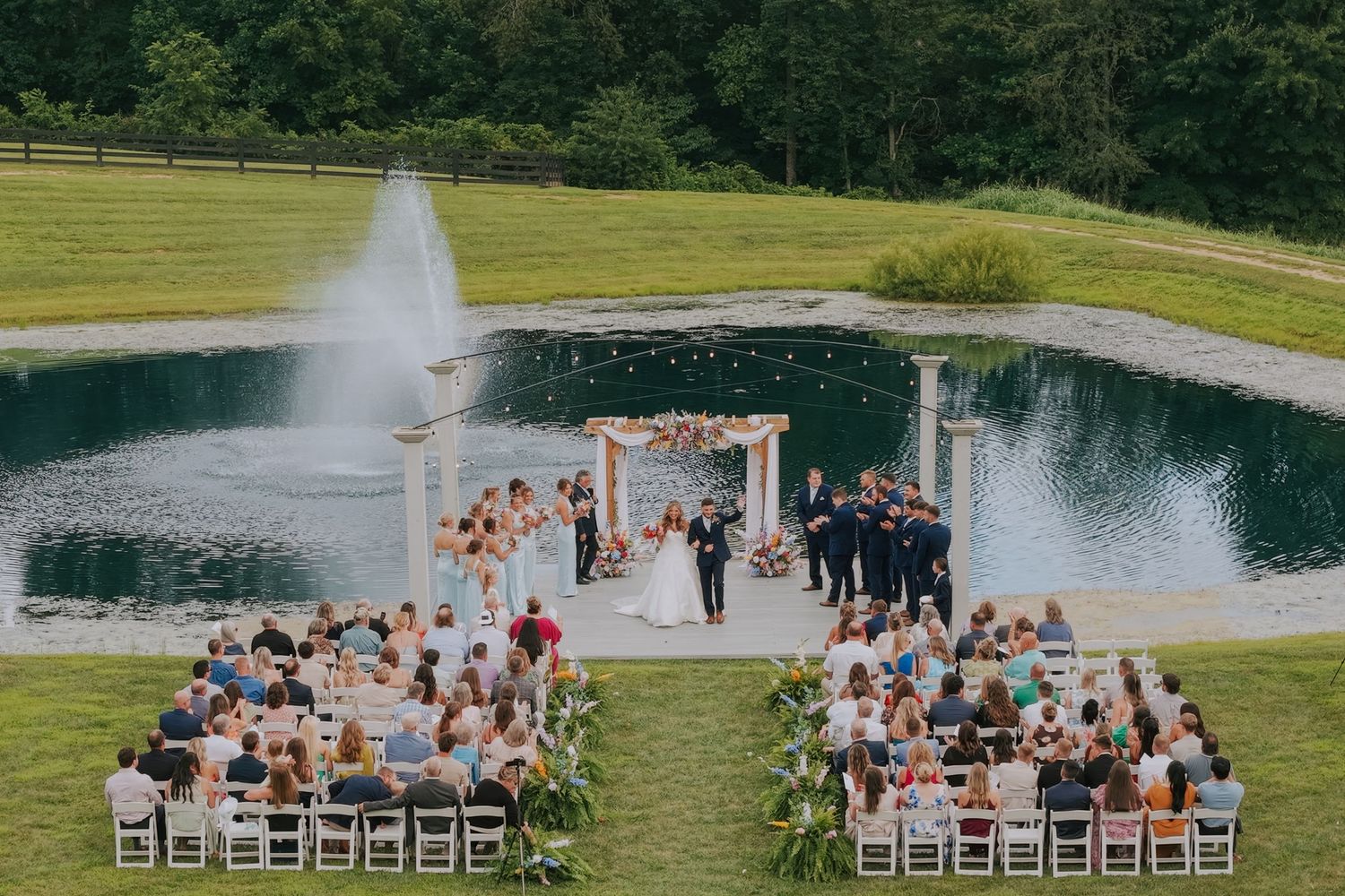 Outdoor wedding ceremony by a pond with fountain, guests seated in white chairs, couple at floral arch altar.