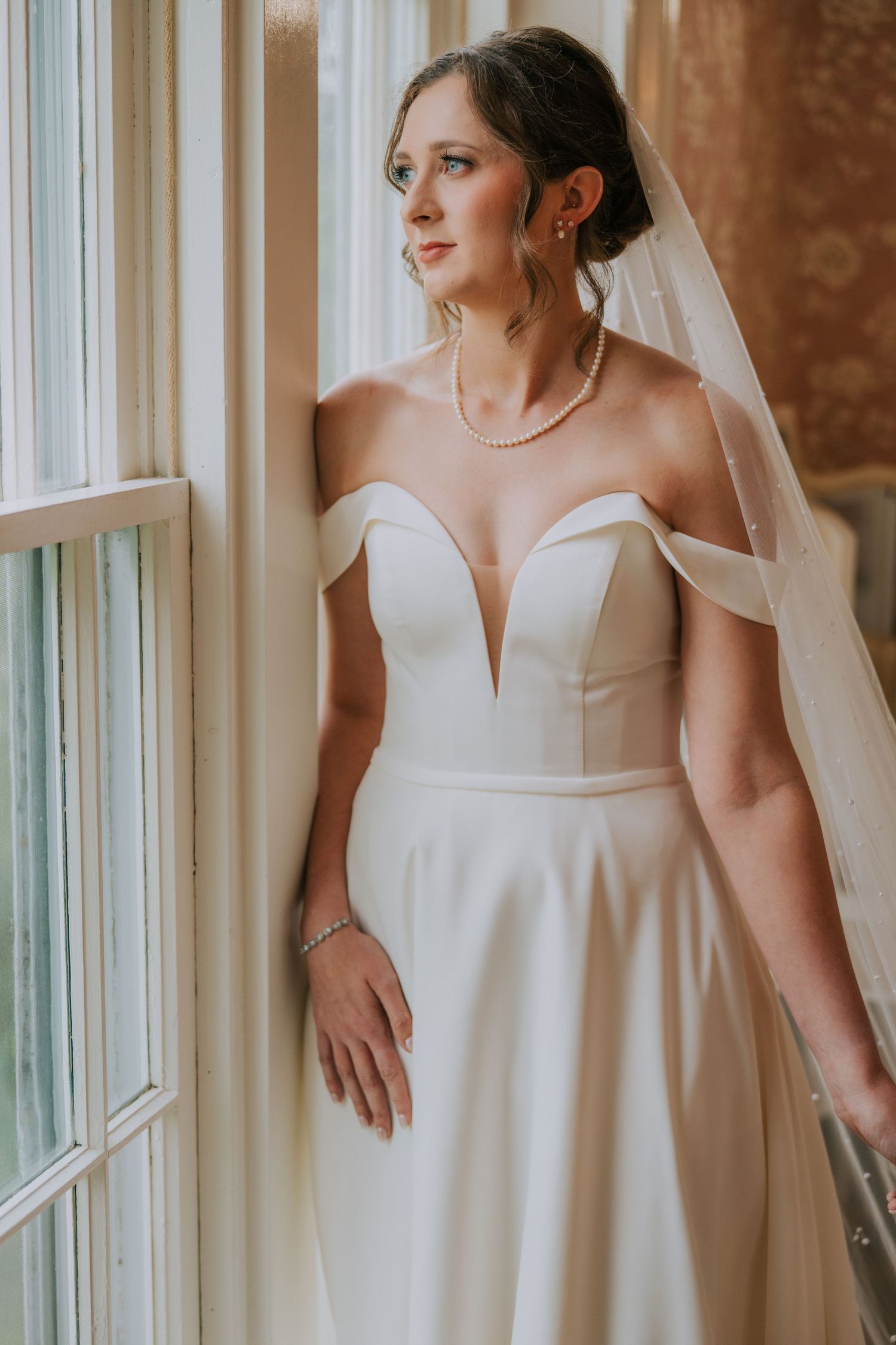 Elegant bridal portrait showing details of a white wedding dress with pearl accessories near sunlit windows.