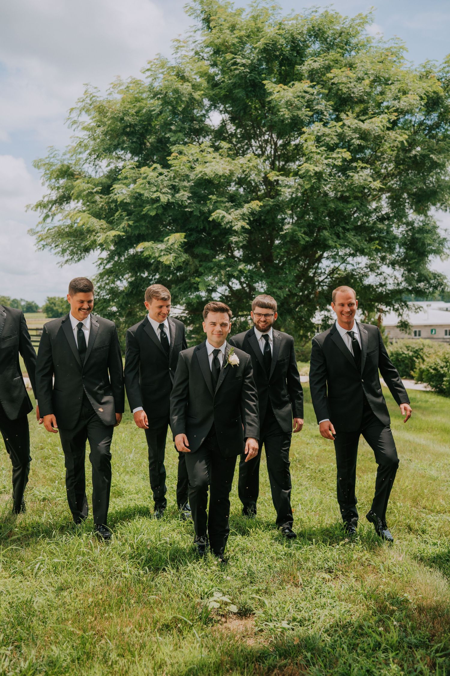 A group of men in matching black suits and ties walk together on green grass with a large tree in the background.