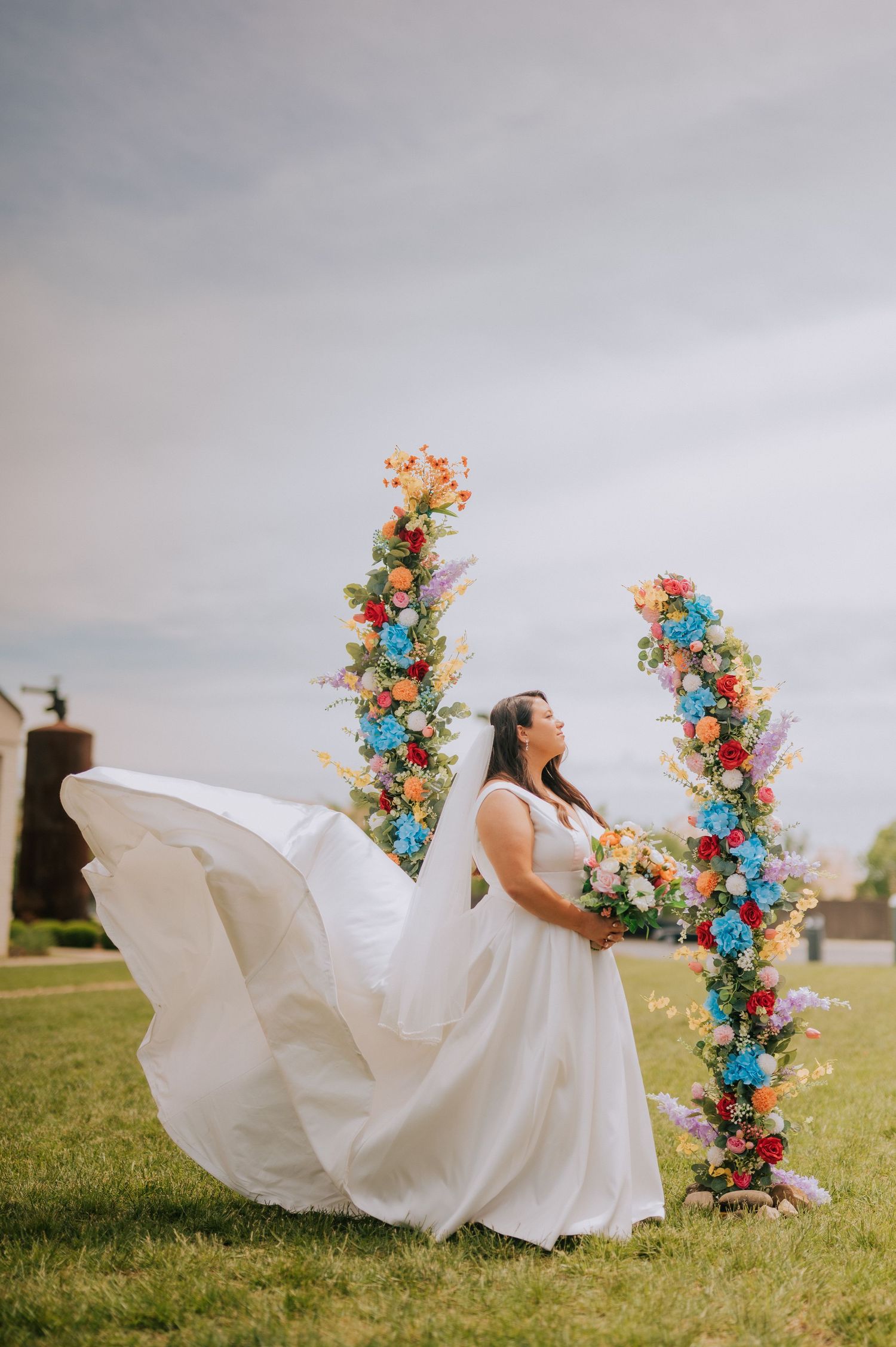 Bride in flowing white dress poses with colorful floral arrangements in outdoor setting.