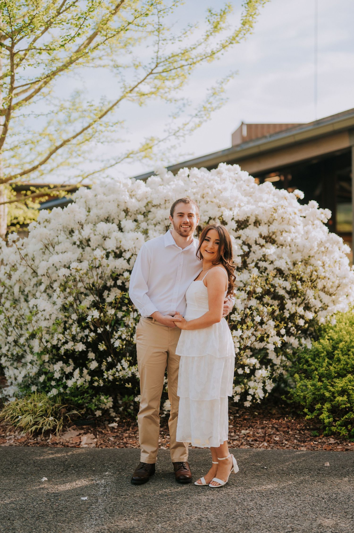 A couple poses together in front of blooming white azalea bushes on a spring day.
