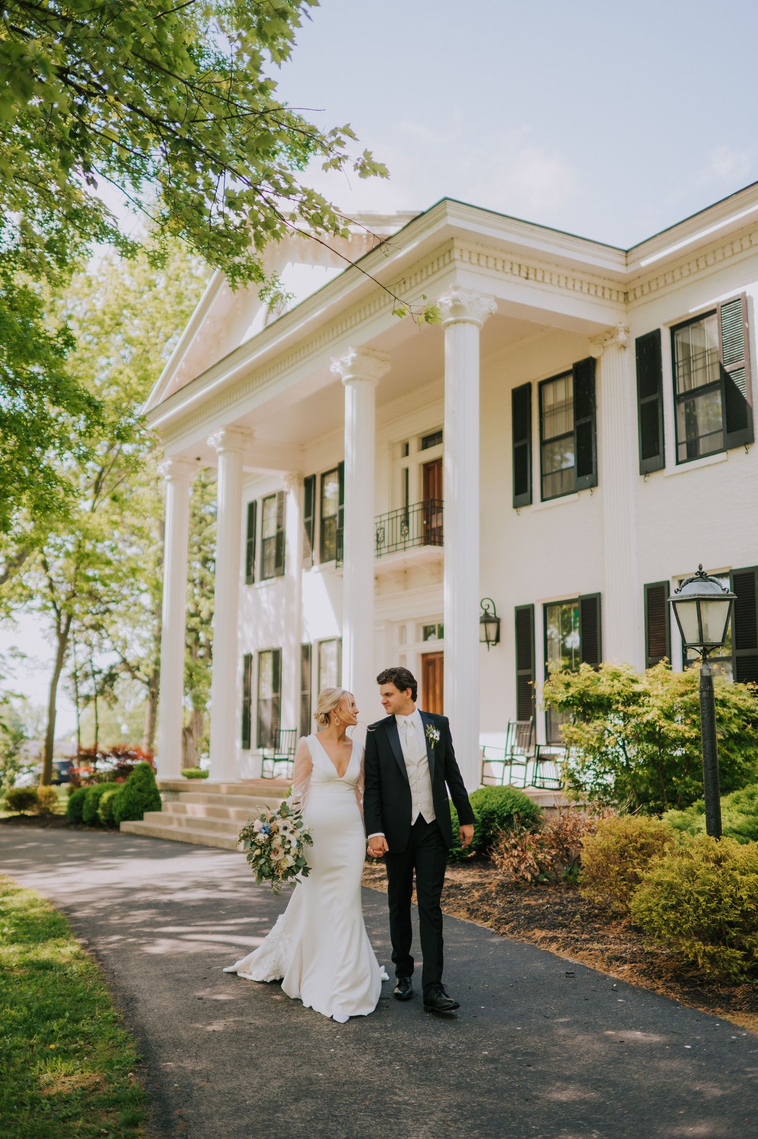 Elegant white antebellum mansion with tall columns and black shutters serves as backdrop for wedding photos.