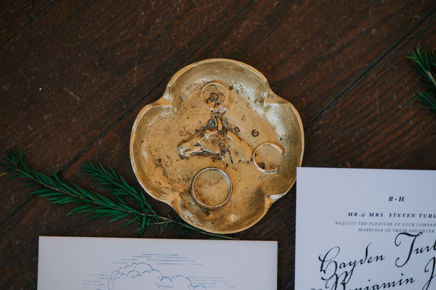 Gold floral-shaped ring dish with wedding rings sits on wooden surface beside paper and greenery.